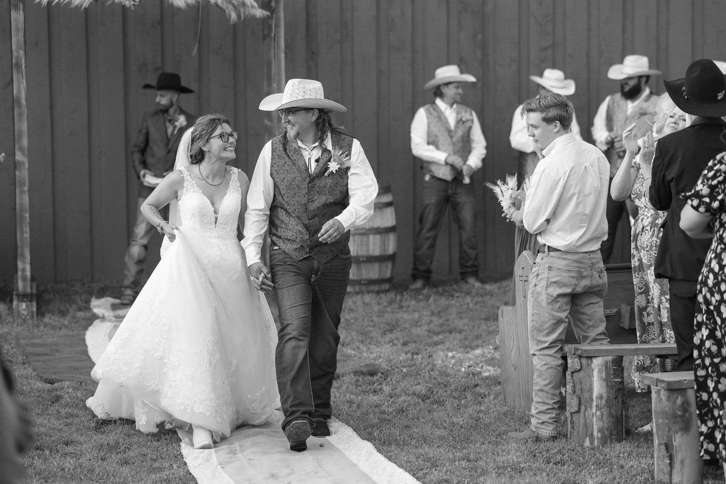 A black and white photo of a wedding ceremony featuring a couple walking down the aisle, holding hands, with guests seated on either side. The bride is in a wedding dress and the groom is in a vest and cowboy hat, indicating a rustic theme.