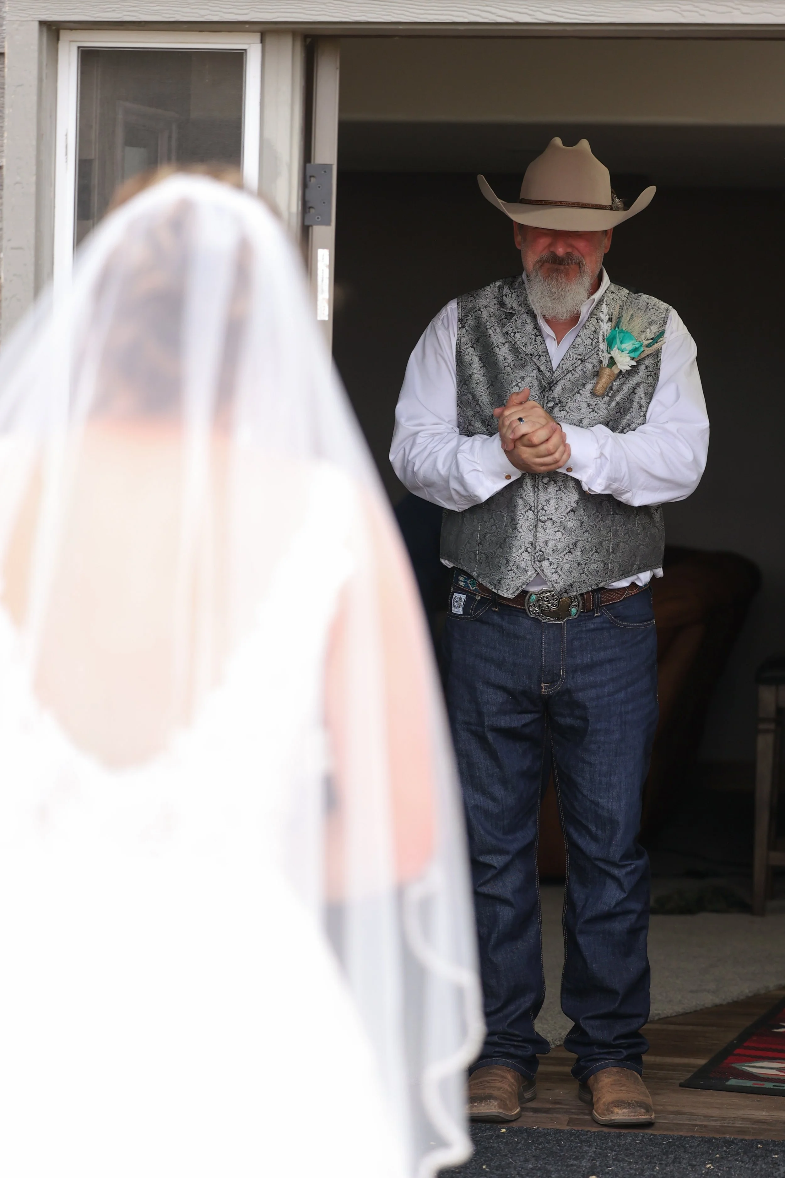 A man dressed in Western attire, including a large cowboy hat, embroidered vest, and jeans, stands with hands clasped in front of him. He has a beard and mustache and is standing at the entrance of a building. The front of a person in a white veil, l