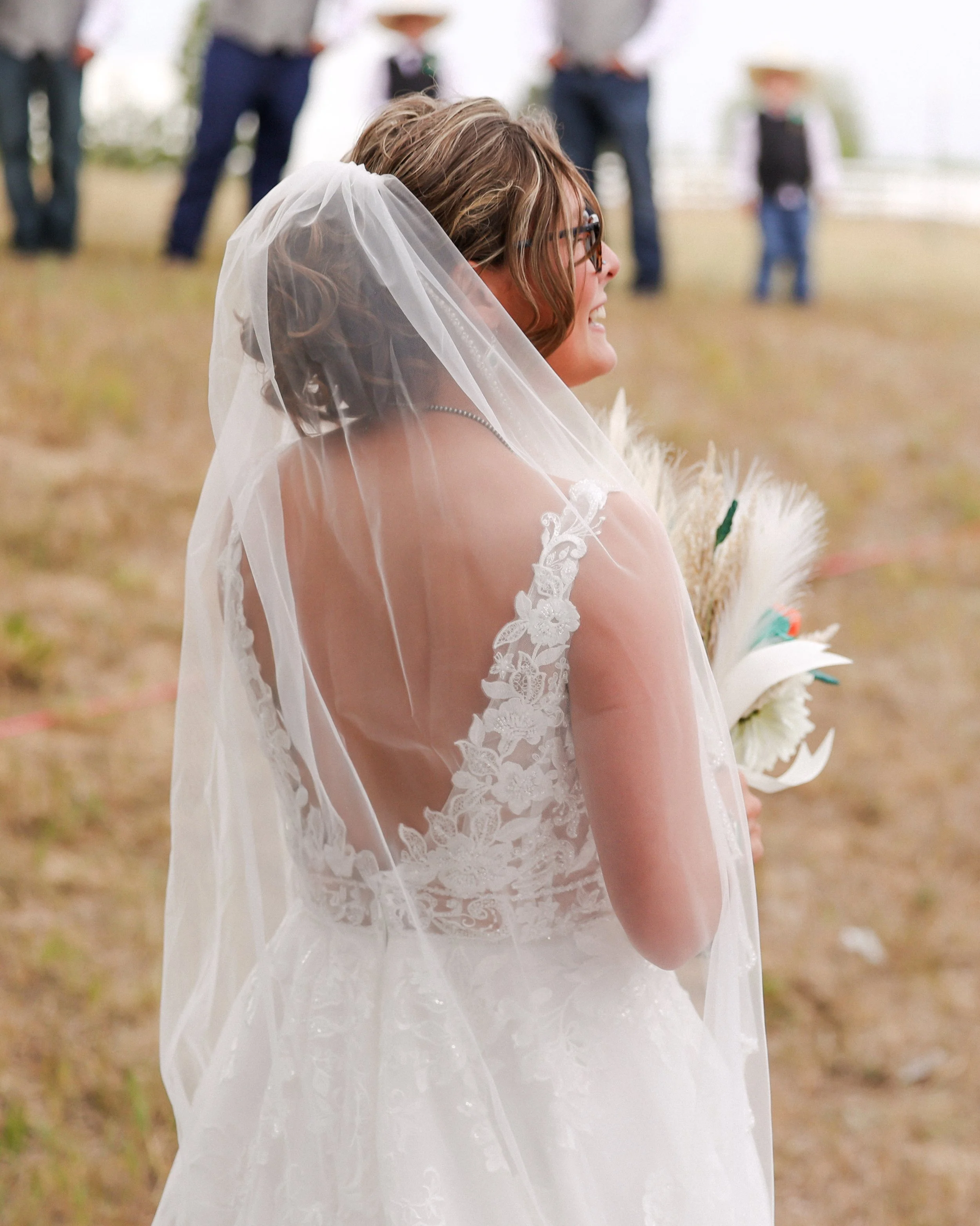 A bride wearing glasses and a white lace wedding dress with a veil, smiling outdoors during her wedding ceremony with guests in the background.