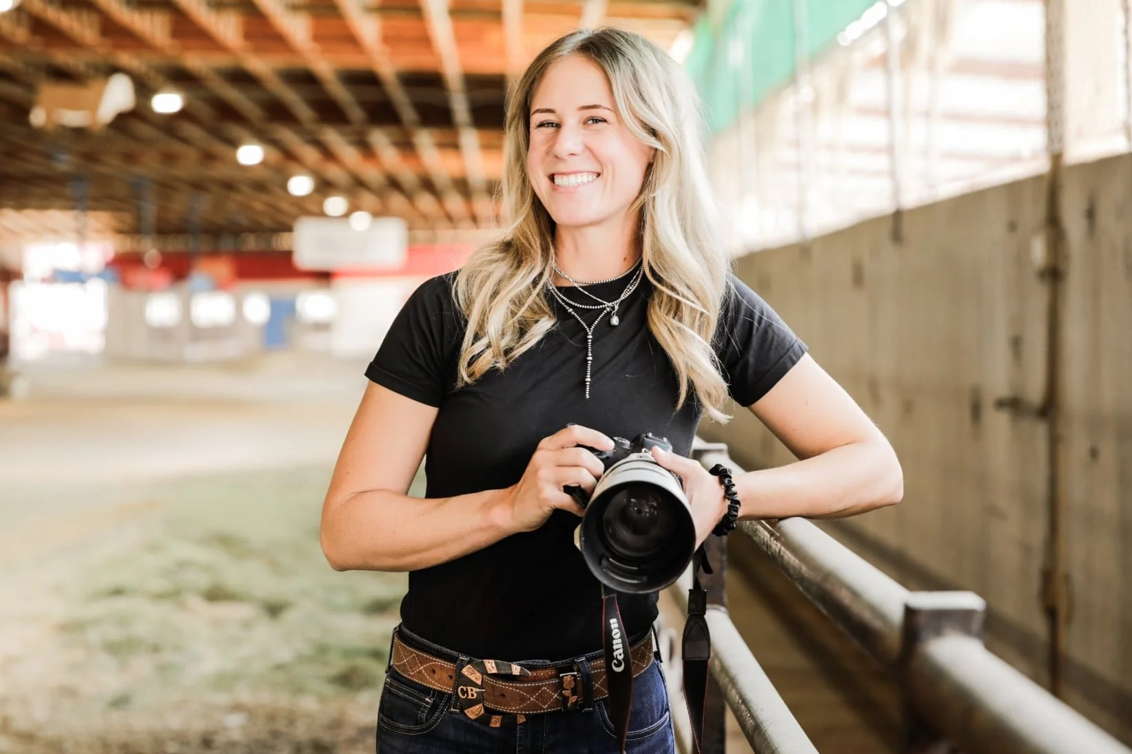 A young woman with blonde hair and a black t-shirt holding a professional camera in an indoor farm or greenhouse setting, smiling at the camera.