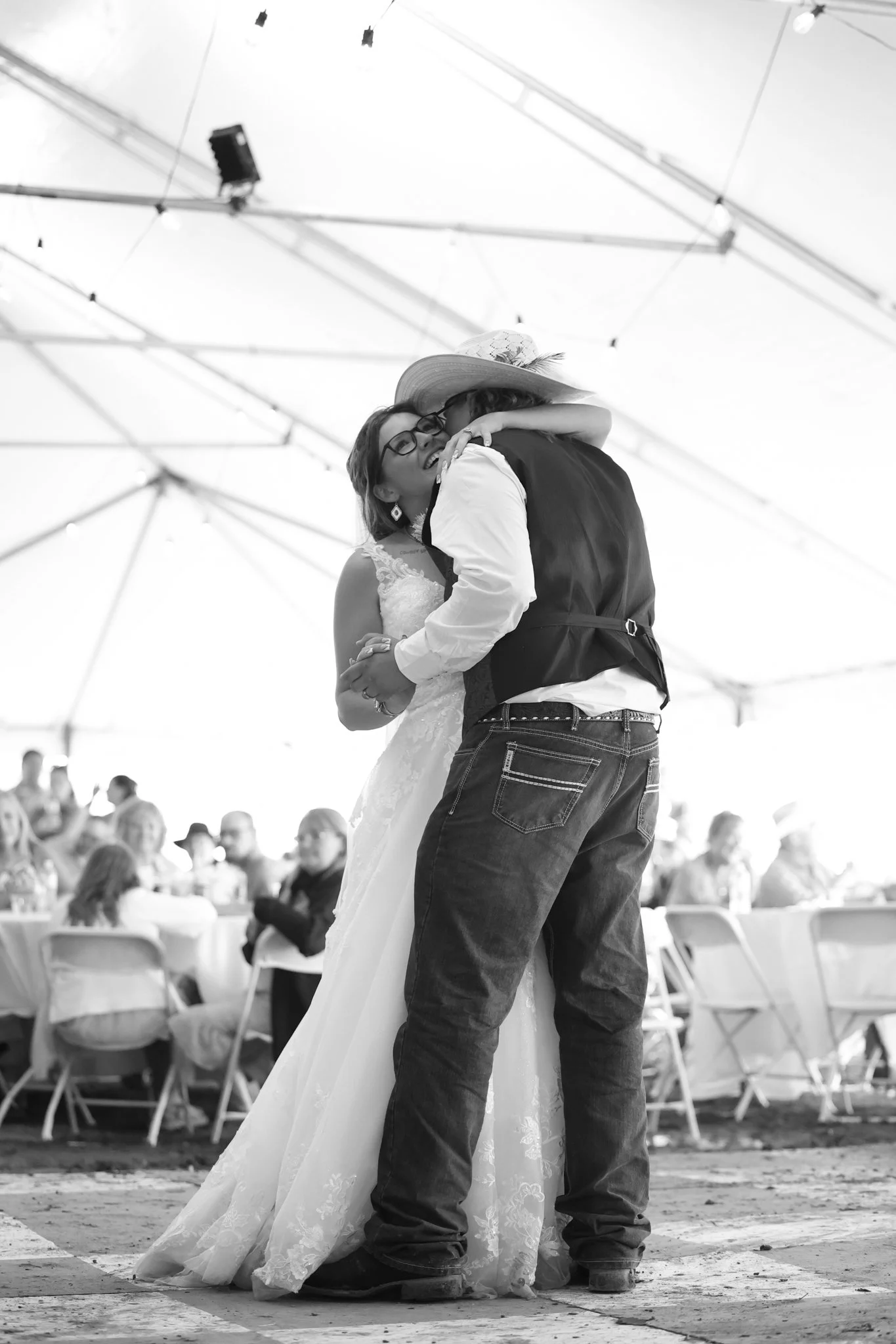 A couple dancing at an outdoor wedding reception, with a woman wearing glasses in a wedding dress and a man in jeans and a vest. Guests sit at tables in the background under a large tent.