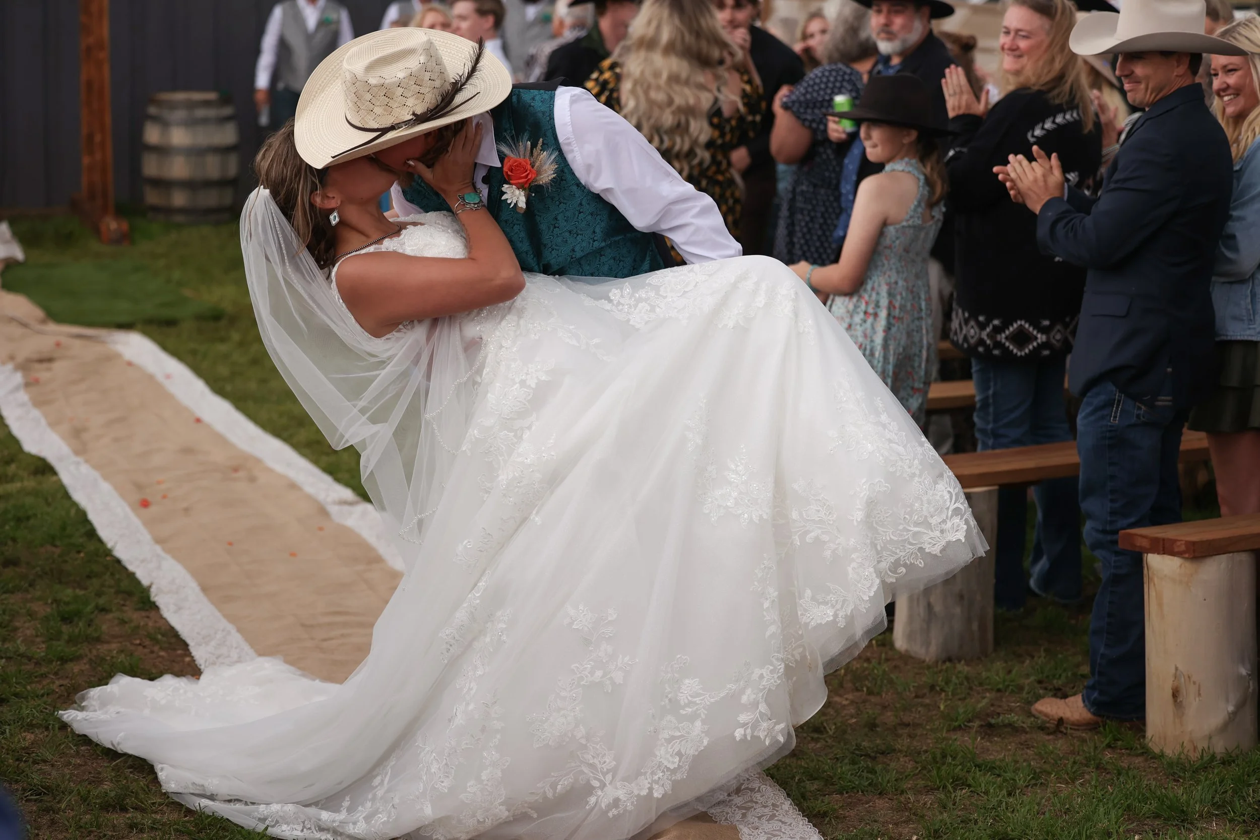 A newlywed couple is sharing a kiss during a wedding ceremony outdoors. The bride is in a white wedding gown and veil, and the groom is in a green vest and cowboy hat. Guests are clapping and smiling in the background.