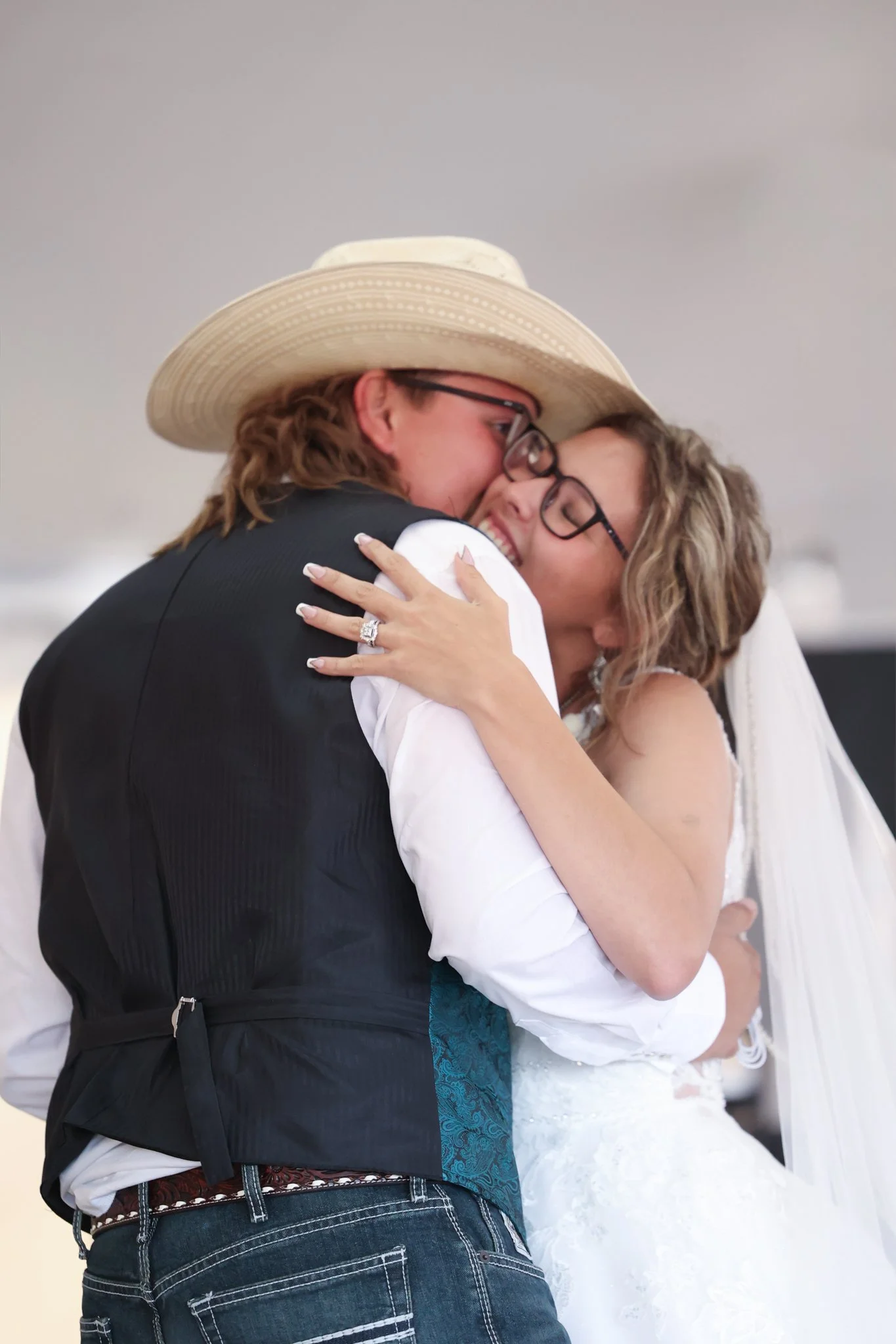 A couple hugging at a wedding, with the woman wearing a white wedding dress and the man wearing a cowboy hat, glasses, and a vest.