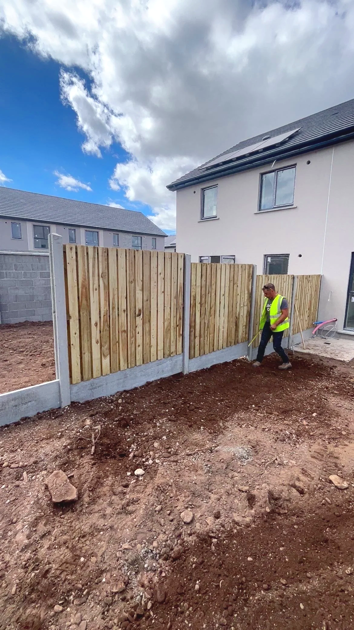 A man in a yellow safety vest is installing a wooden fence on a construction site with an unfinished ground surface, in front of residential buildings under a partly cloudy sky.