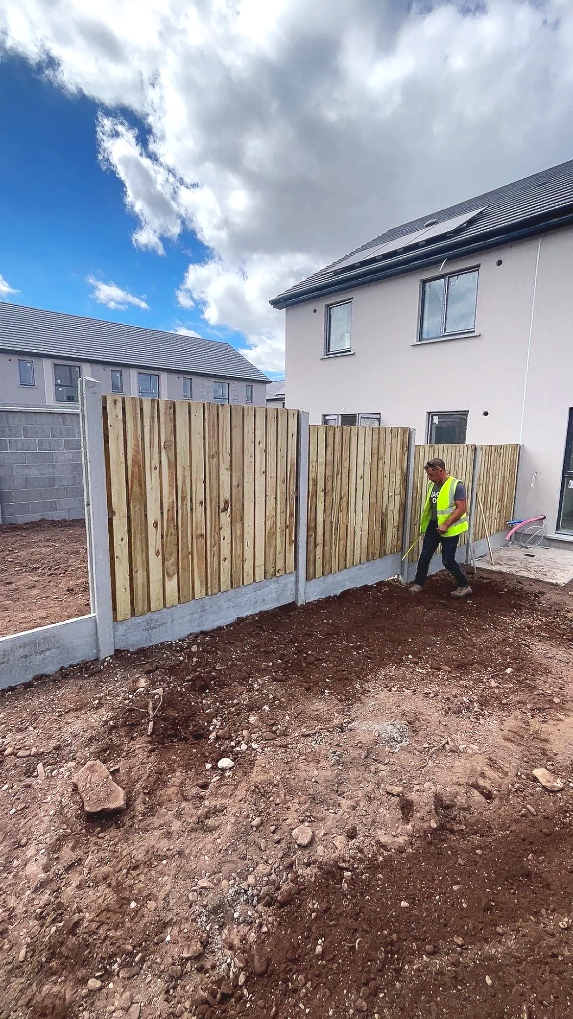 A person in a yellow safety vest working on a wooden fence in a backyard with unfinished ground, under a cloudy sky.