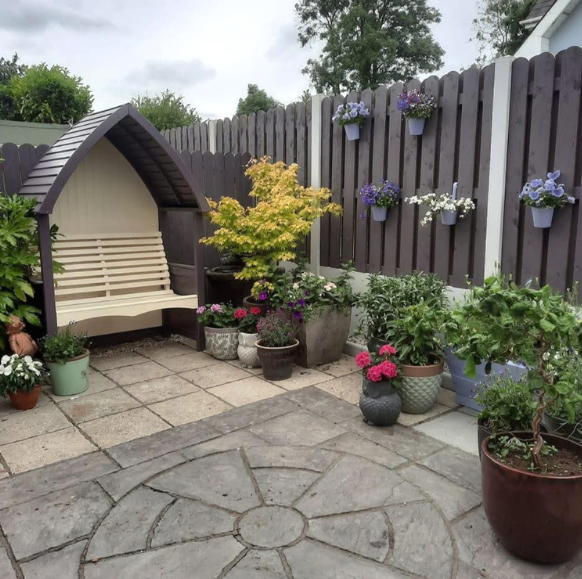 Backyard garden with potted plants, hanging flowers on a wooden fence, and a beige bench under a small garden house in Villerstown County Waterford.