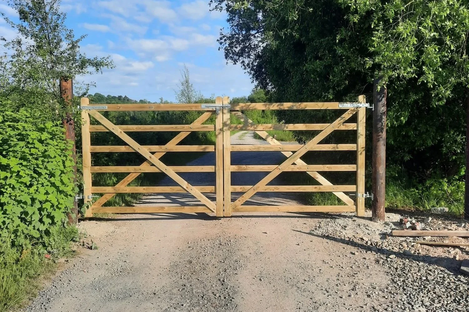 Wooden gate with diagonal cross bracing, leading to a gravel path beside a waterway, surrounded by green trees and shrubs under a partly cloudy sky.