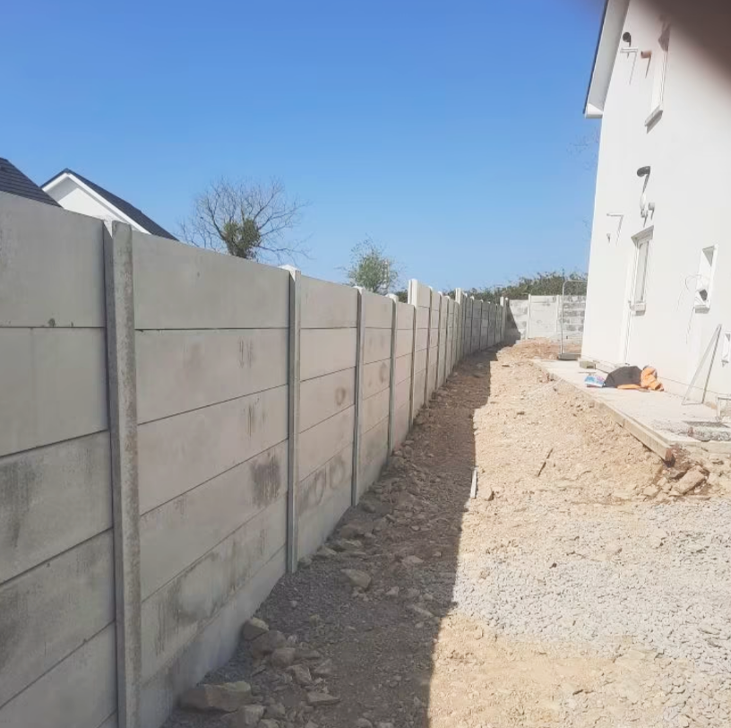 A newly built concrete fence runs alongside a dirt and gravel pathway next to a white building under construction, with a clear blue sky overhead.