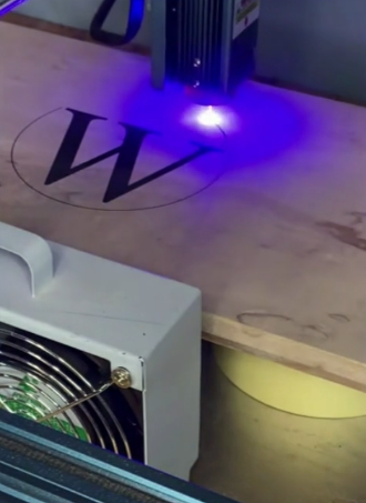 A laser engraver engraving a large letter 'W' on a wooden surface with a circular outline around it.