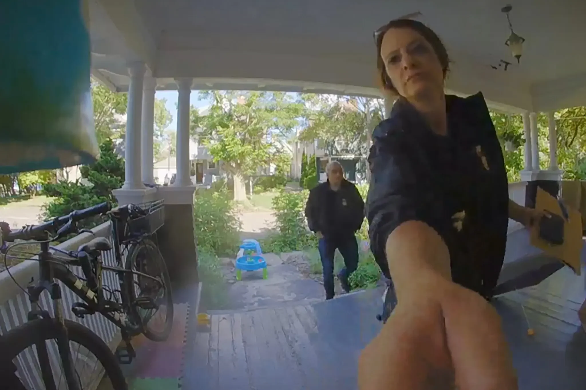 Two women are standing on a porch with white columns and greenery in the background. One woman is closer to the camera, wearing a black top and glasses, looking slightly to the side. The other woman is in the background, wearing sunglasses and a black jacket. Several bicycles are parked along the porch railing, and a small blue and green children's toy is on the ground.