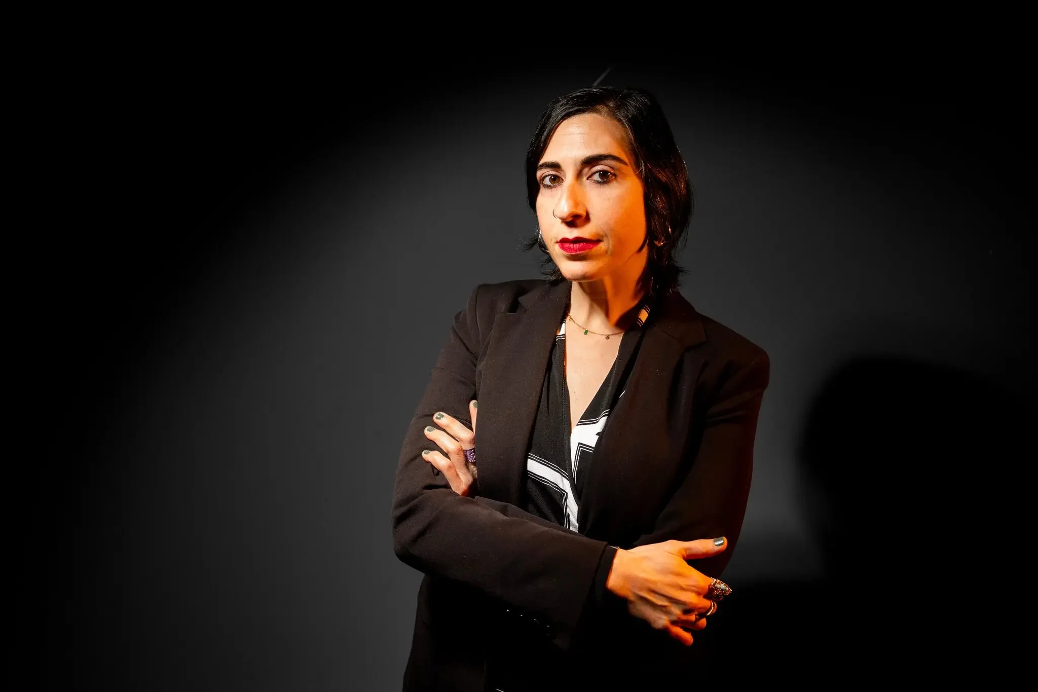 A woman with short black hair, wearing a black blazer and a striped shirt, standing against a dark background. She is crossing her arms and looking at the camera with a confident expression.