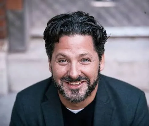 A smiling man with dark hair and a beard, wearing a dark blazer, sitting outdoors against a background of stairs.