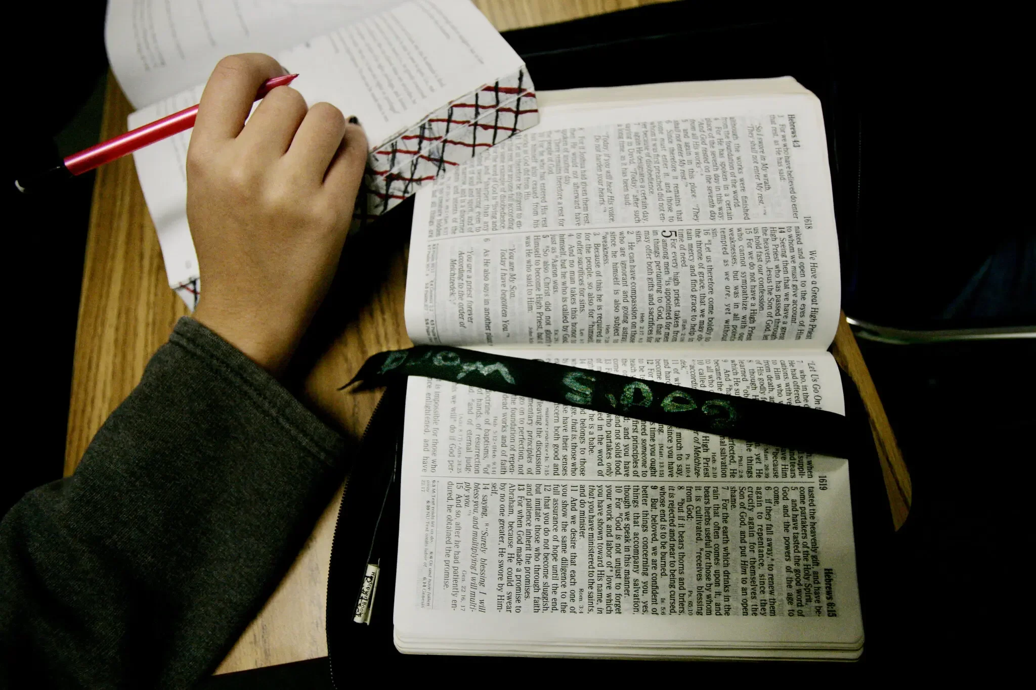 A person's hand holding a red pen, writing on a paper placed over an open book on a wooden table.