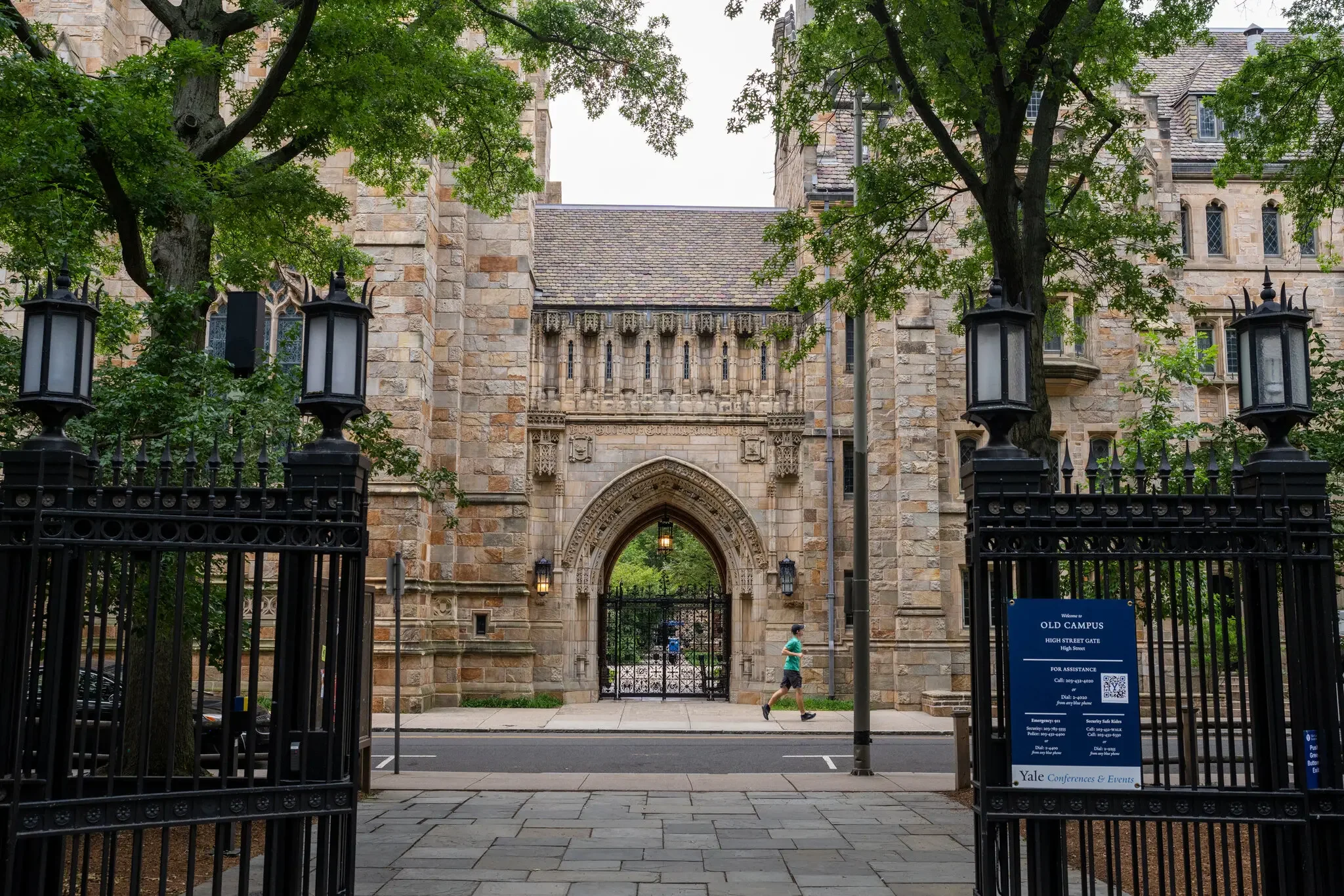 Stone gate entrance to Yale campus with decorative arches, wrought iron fencing, and a blue informational sign. Green trees partially frame the scene, and two people walk along the sidewalk.