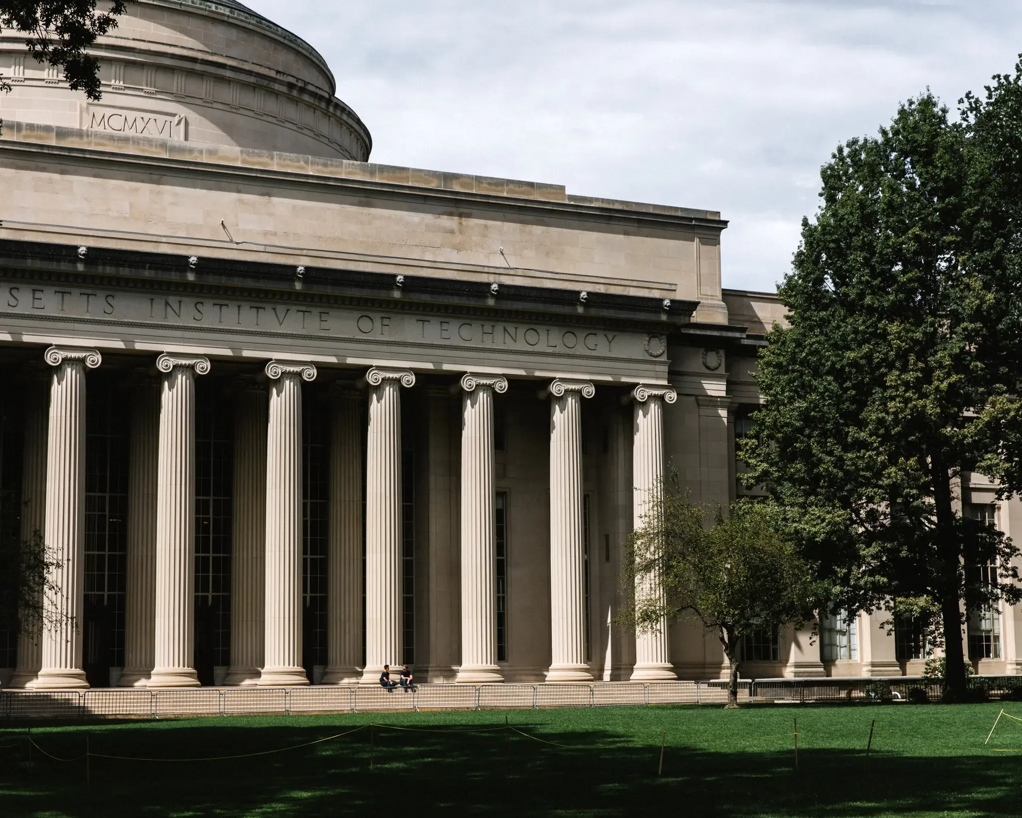 The image shows a large classical-style building with tall columns and a rounded facade. There are four people sitting on the steps in front of the building, and trees with green leaves are seen on the right side. The sky above is mostly cloudy.