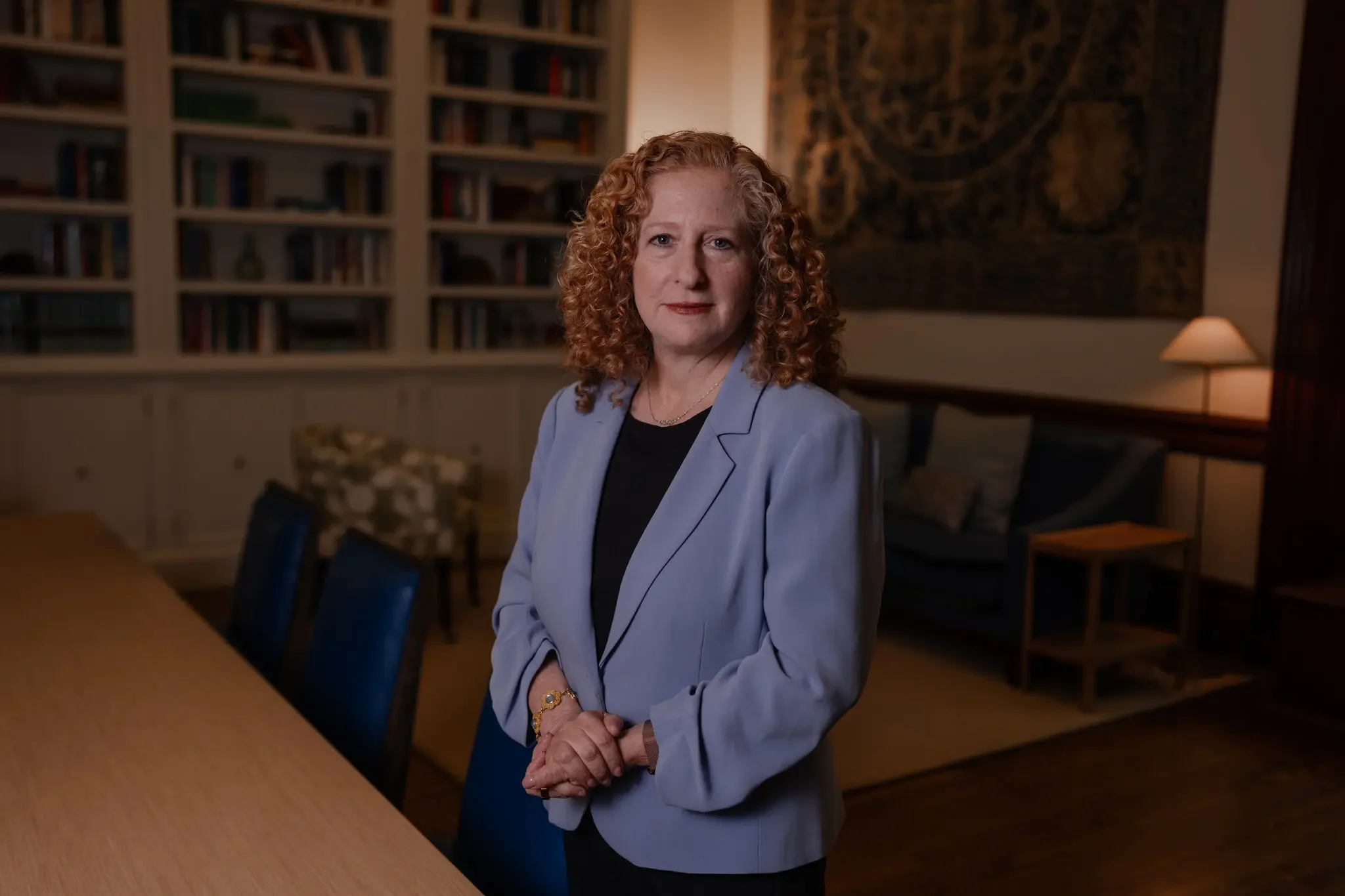 A woman with curly red hair, wearing a gray blazer and black top, sits at a conference table in a room with a bookshelf and a tapestry on the wall behind her.