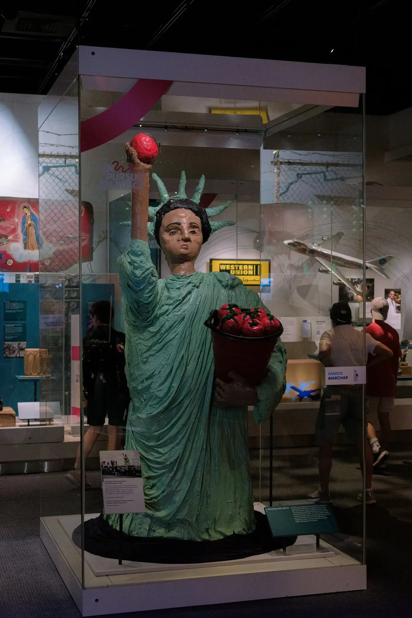 Sculpture of the Statue of Liberty inside a glass display case at a museum, with its right arm raised holding a torch topped with a red object and a bouquet of red flowers in front.