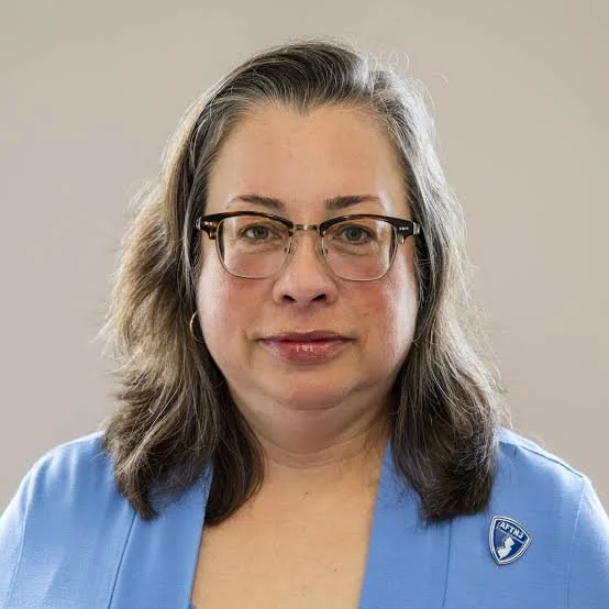 A woman with shoulder-length gray hair, glasses, and hoop earrings, wearing a blue blazer with an emblem on it, standing against a plain background.