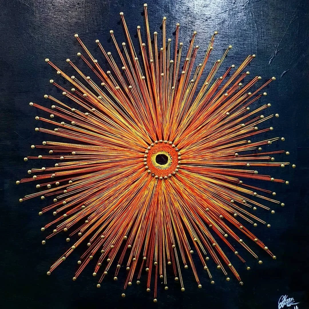 A radial artwork made with orange, yellow, and brown colored sticks and small rounded beads at the ends, arranged in a starburst pattern on a dark background.