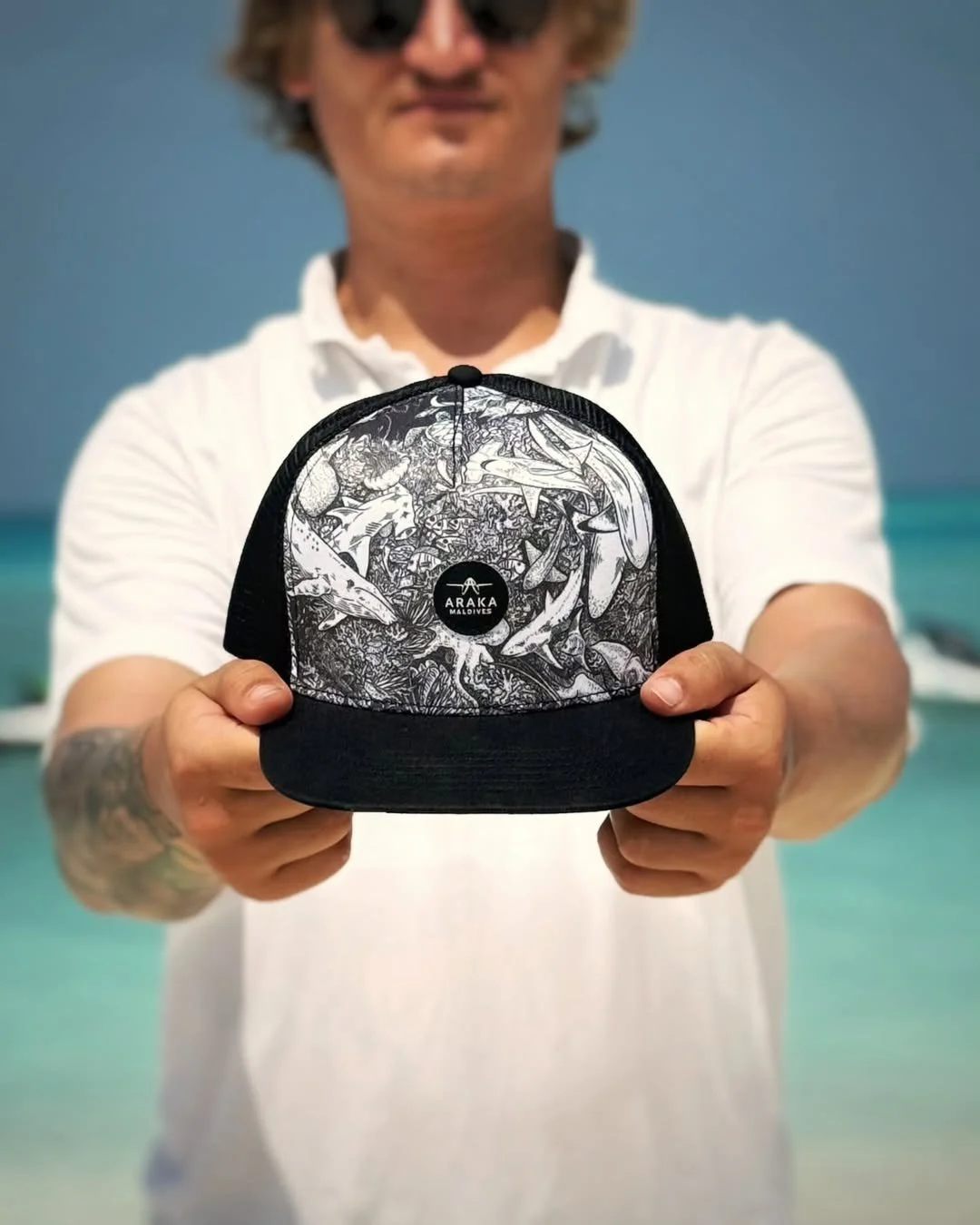 A man holding a black and white baseball cap with marine life illustrations, against a beach background.
