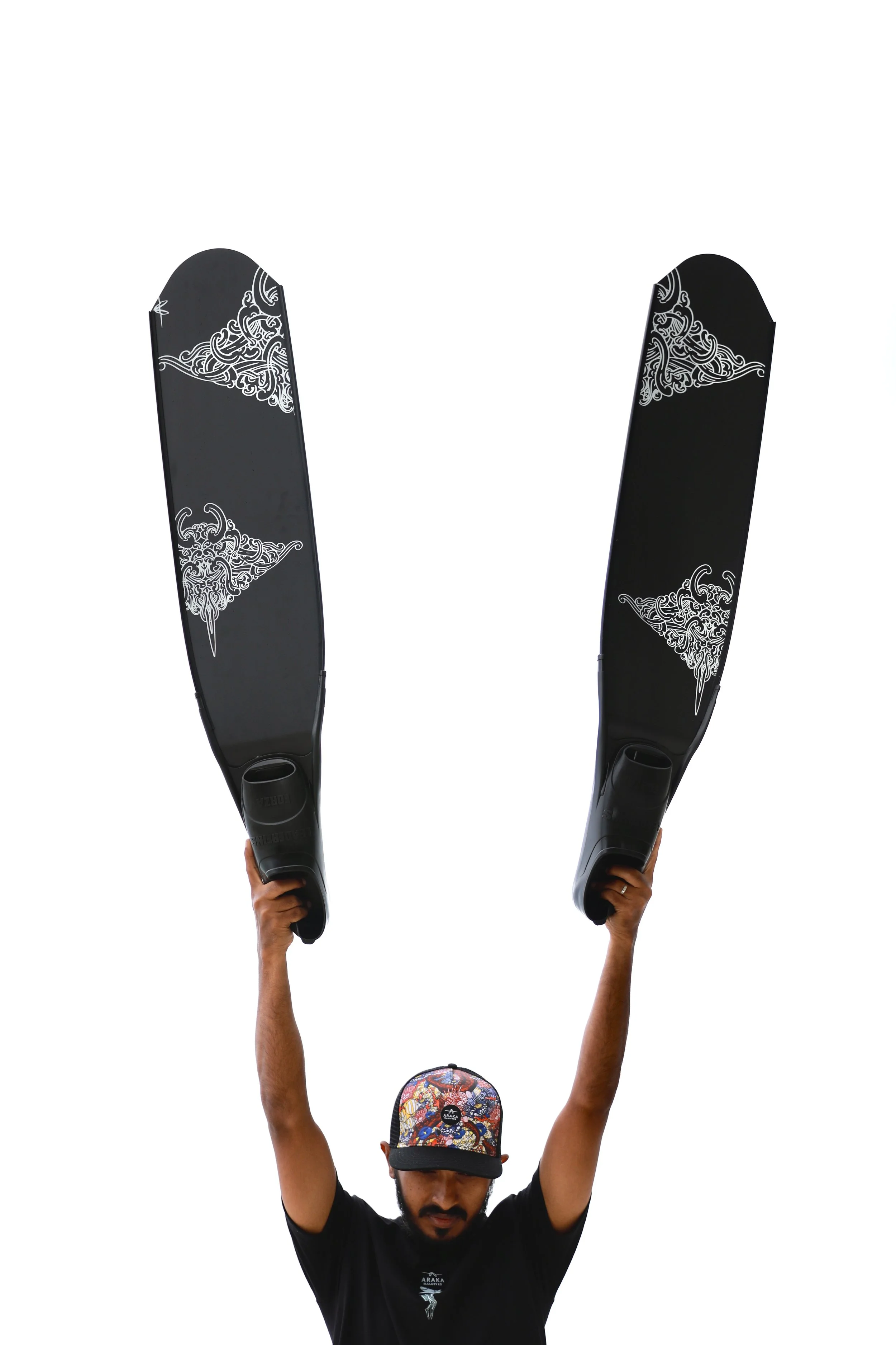 A man wearing a colorful cap and black T-shirt holding two large black surfboards with white decorative designs, raised above his head against a white background.