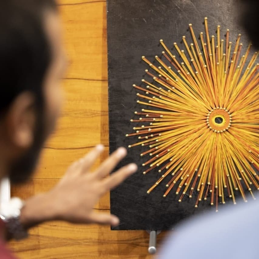 People observing a yellow and orange sunburst wall art sculpture made of wooden sticks and round knobs.