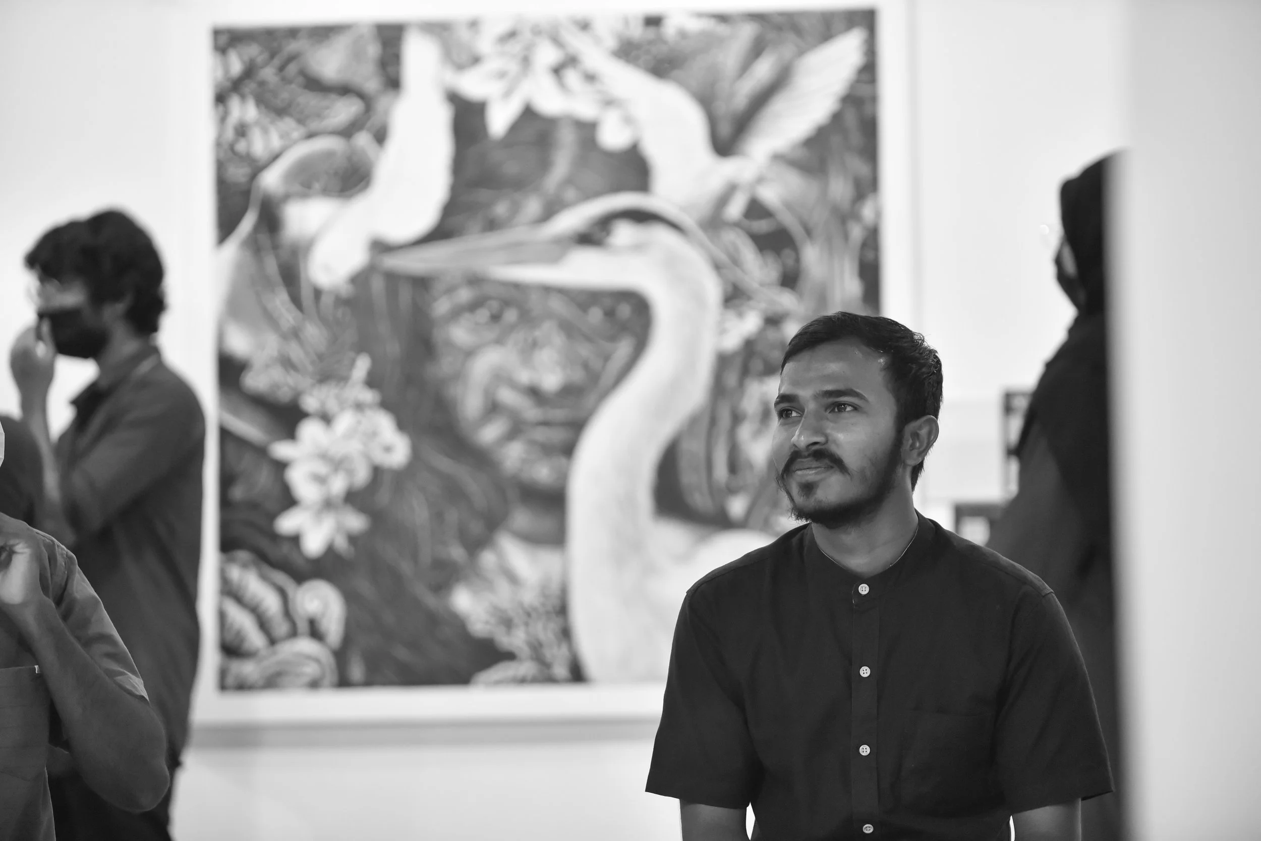 A black and white photo of a man with a beard and mustache looking slightly to the right, in an art gallery with a large painting of a bird in the background. There are other people in the background, one with glasses and a beard, and another with a hijab, all observing the artwork.