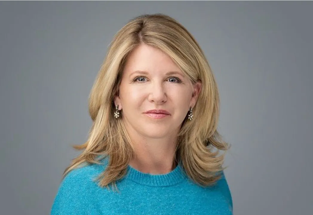 A woman with shoulder-length blonde hair, wearing a blue sweater and pearl earrings, against a gray background.