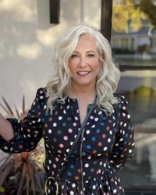 A woman with long, curly gray hair smiling outdoors, wearing a black dress with colorful polka dots, in front of a window with greenery and trees visible.