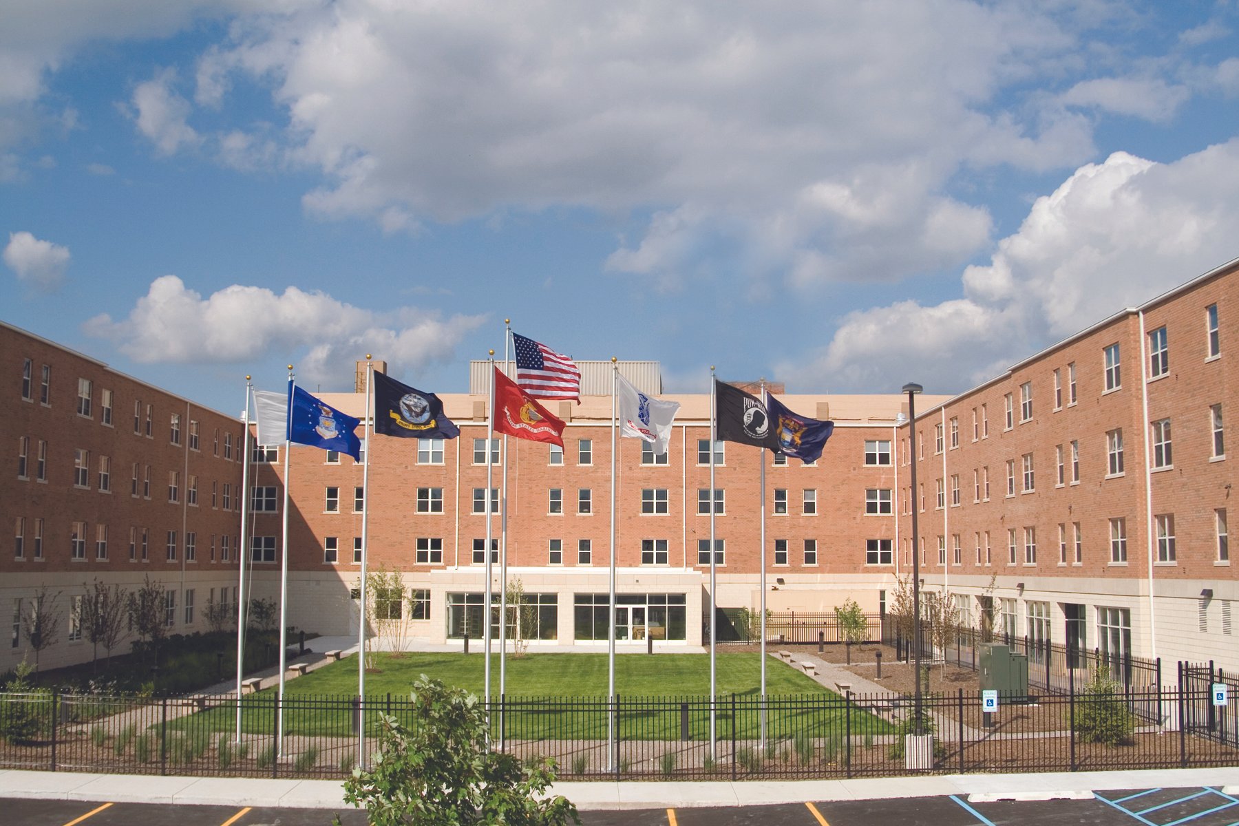 A courtyard with grass and small trees enclosed by a black metal fence, in front of a large brick building with multiple windows. Eight flags, including the United States flag, are flying on poles in the center of the courtyard against a partly cloudy sky.