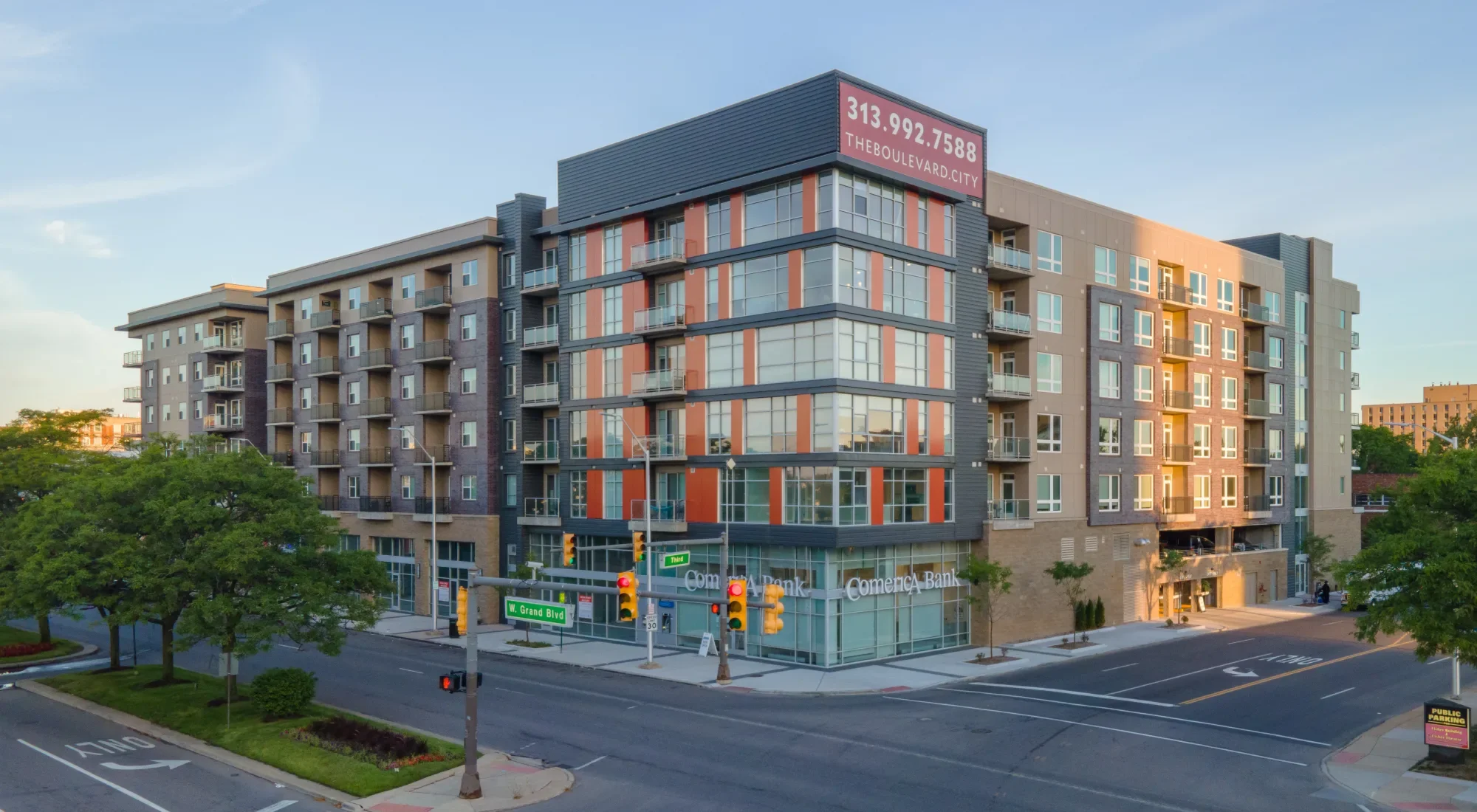 A modern multi-story building with commercial space on the ground floor and residential units above, located at the corner of an intersection on West Grand Blvd, with street signs, traffic lights, trees, and a blue sky.