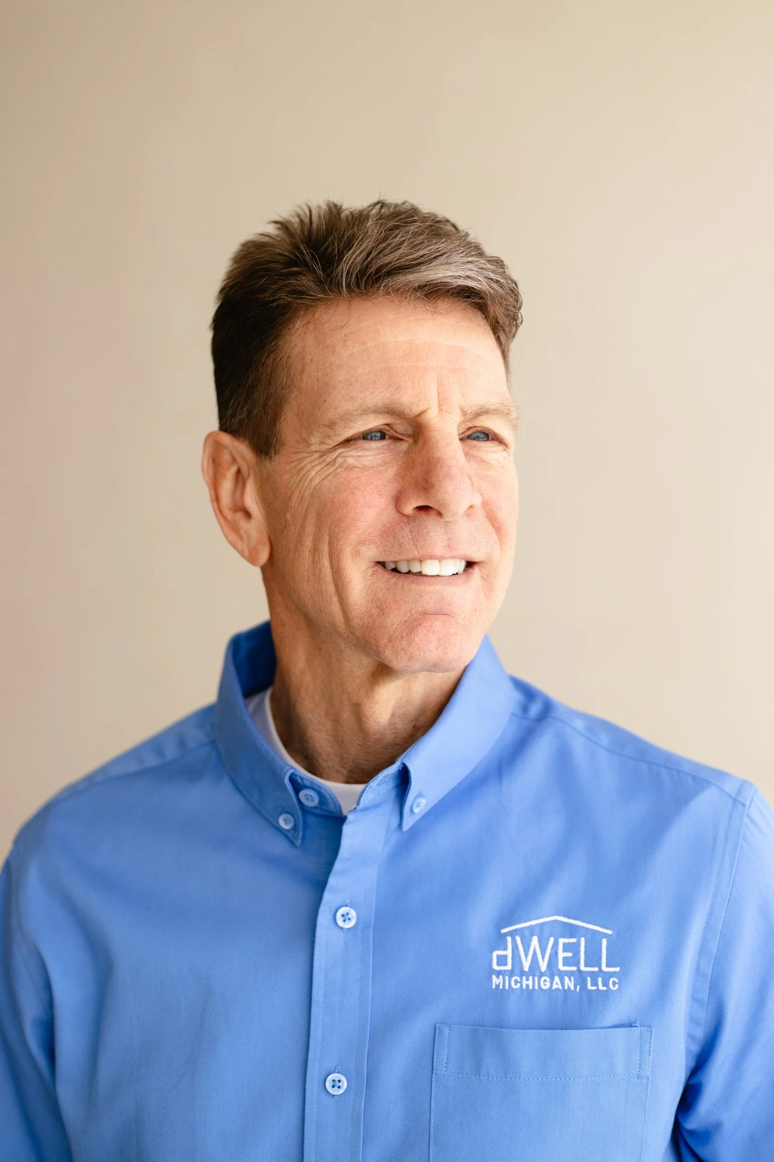 A middle-aged man with light skin, brown hair, and blue eyes, smiling slightly while looking off to the side. He is wearing a blue button-up shirt with a company logo that reads "Dwell Michigan, LLC" embroidered on the chest. The background is plain and light-colored.