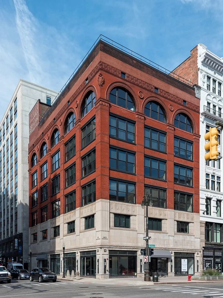 A multi-story brick building with large window arches on top, located at a street corner with cars and a traffic light in view.