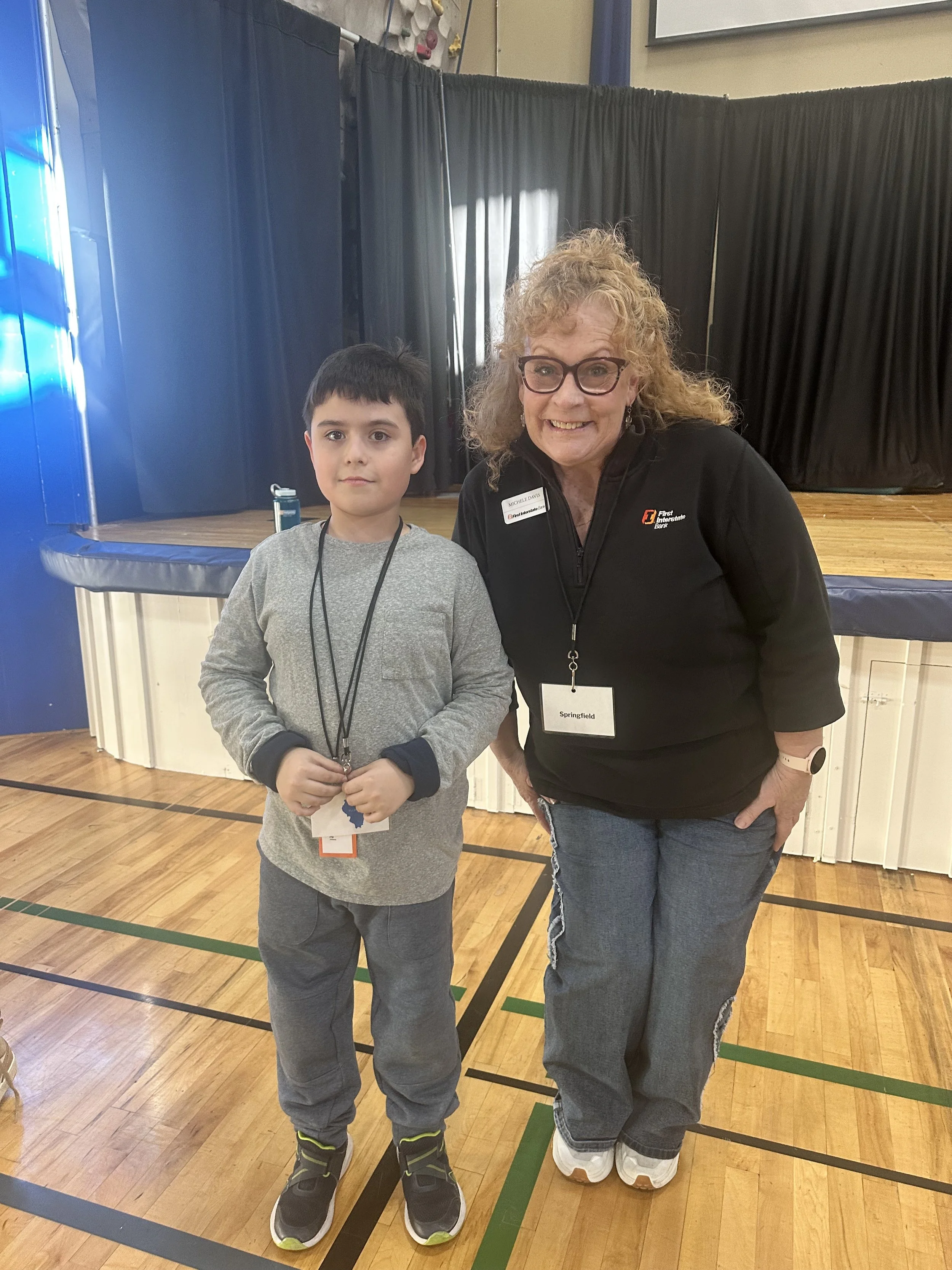 A woman and a boy standing side by side indoors, smiling for the camera. The woman has curly blonde hair, glasses, and is wearing a black zip-up jacket with a name tag and black pants. They are on a wooden gymnasium floor, with black curtains and sta