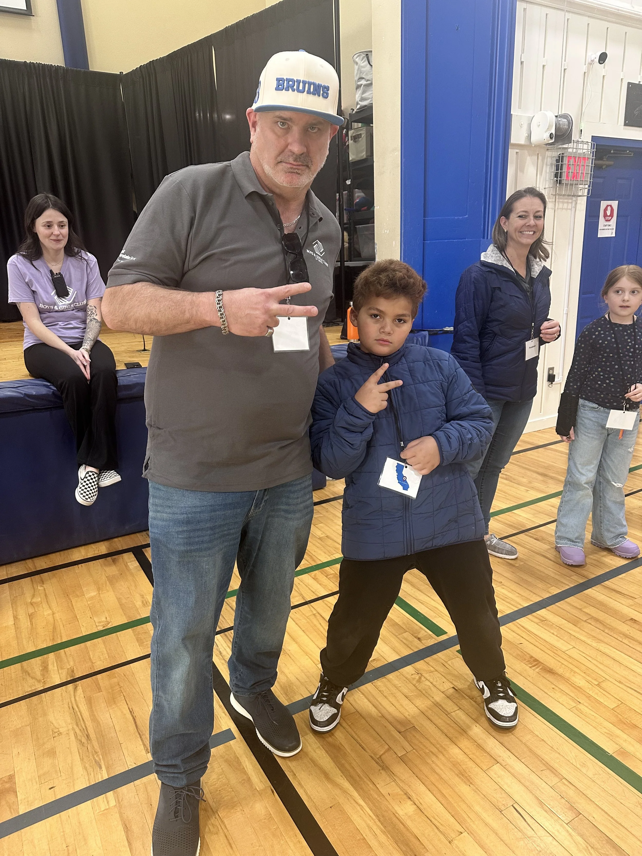 A group of people in a gymnasium, with two men and three children. The man in the foreground is wearing a gray polo shirt, jeans, and a white cap with blue lettering that says 'Bruins.' He is making a peace sign. The boy next to him is in a blue jack