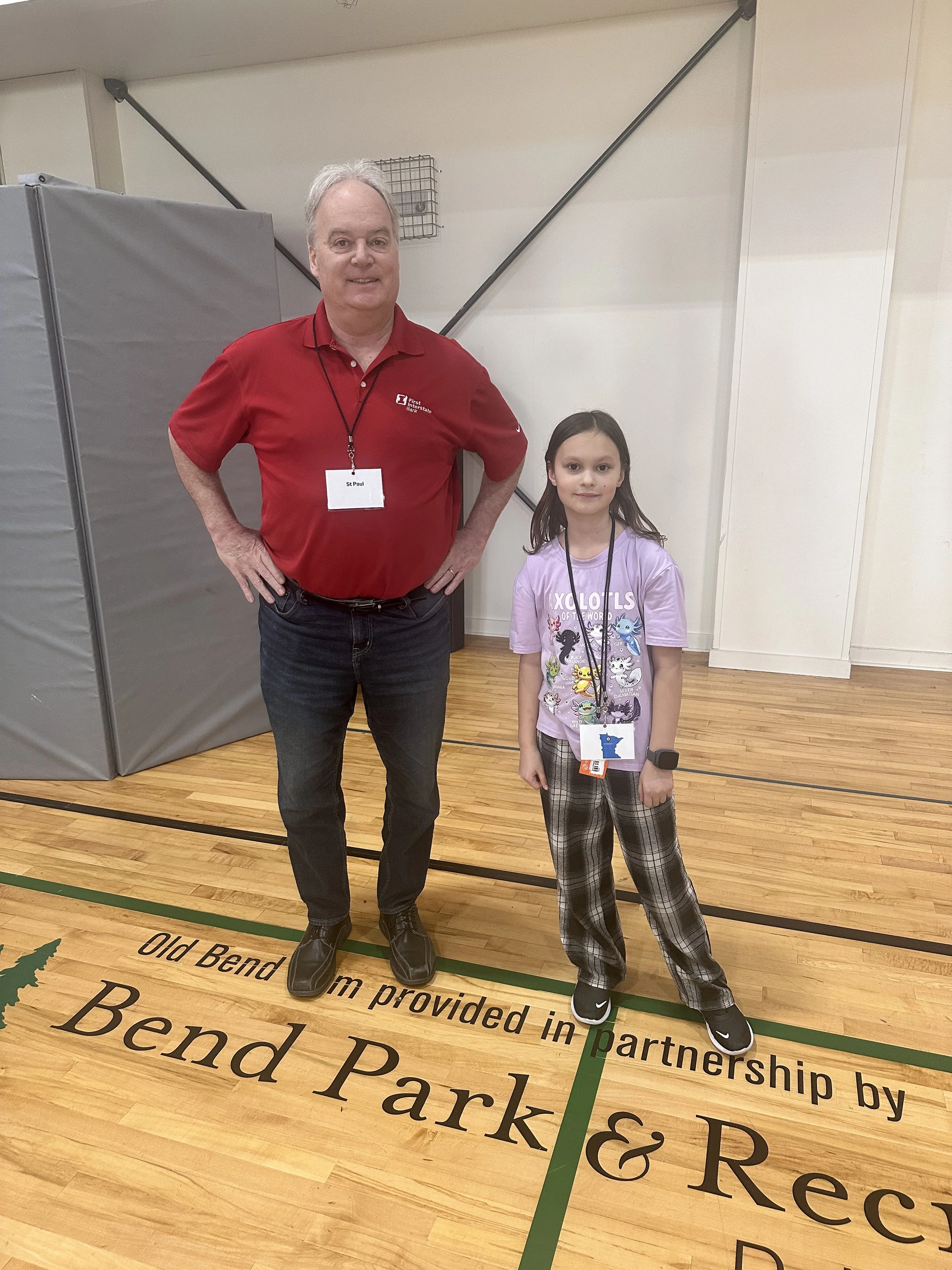 A man and a young girl stand together in a gymnasium, both wearing name badges and casual clothing. The man is dressed in a red polo shirt and jeans, and the girl is wearing a T-shirt with cartoon characters and plaid pants. They are smiling at the c