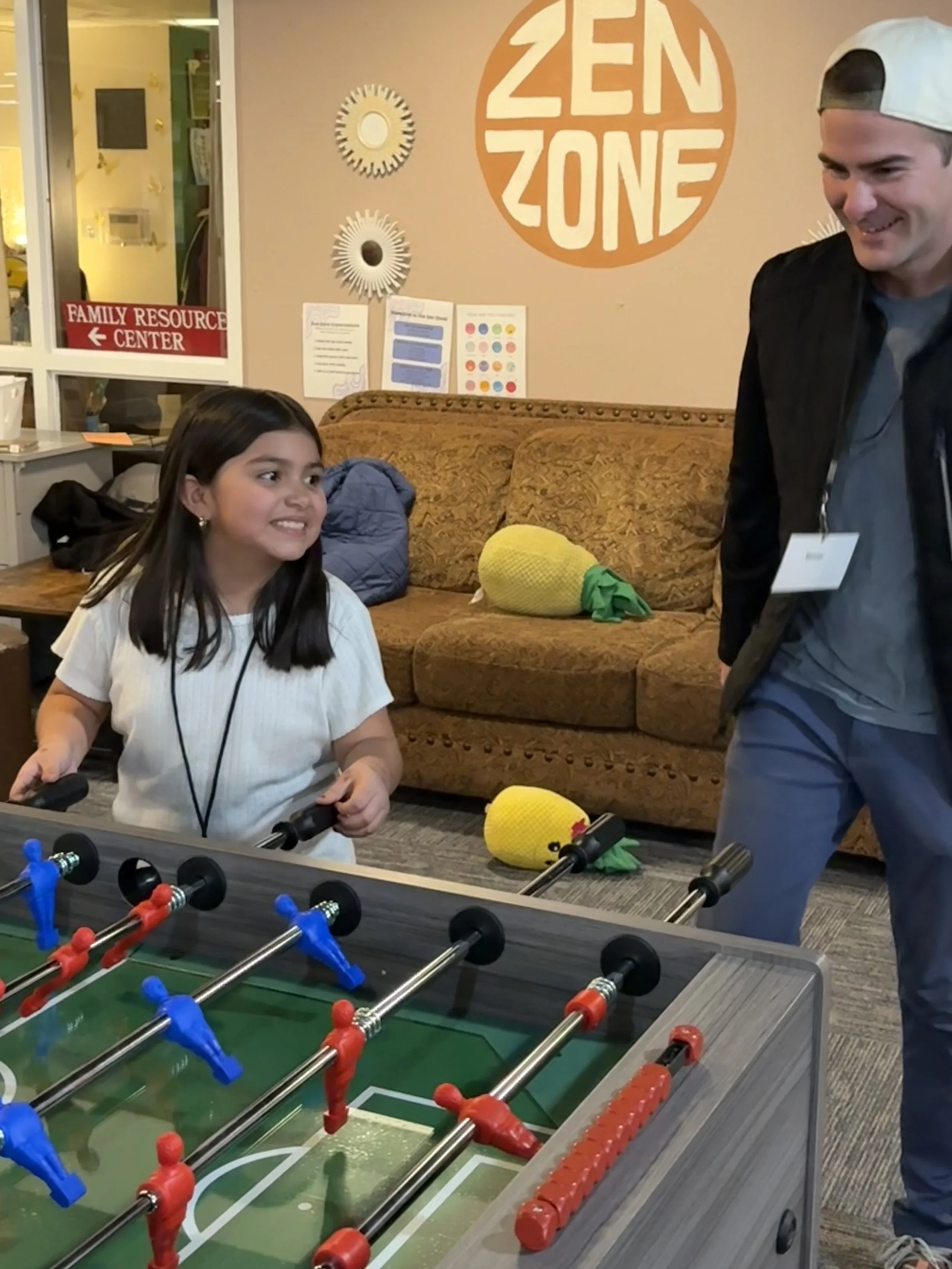 A young girl playing foosball in a room with a couch and wall decorations, while a man in a cap and jacket smiles at her.