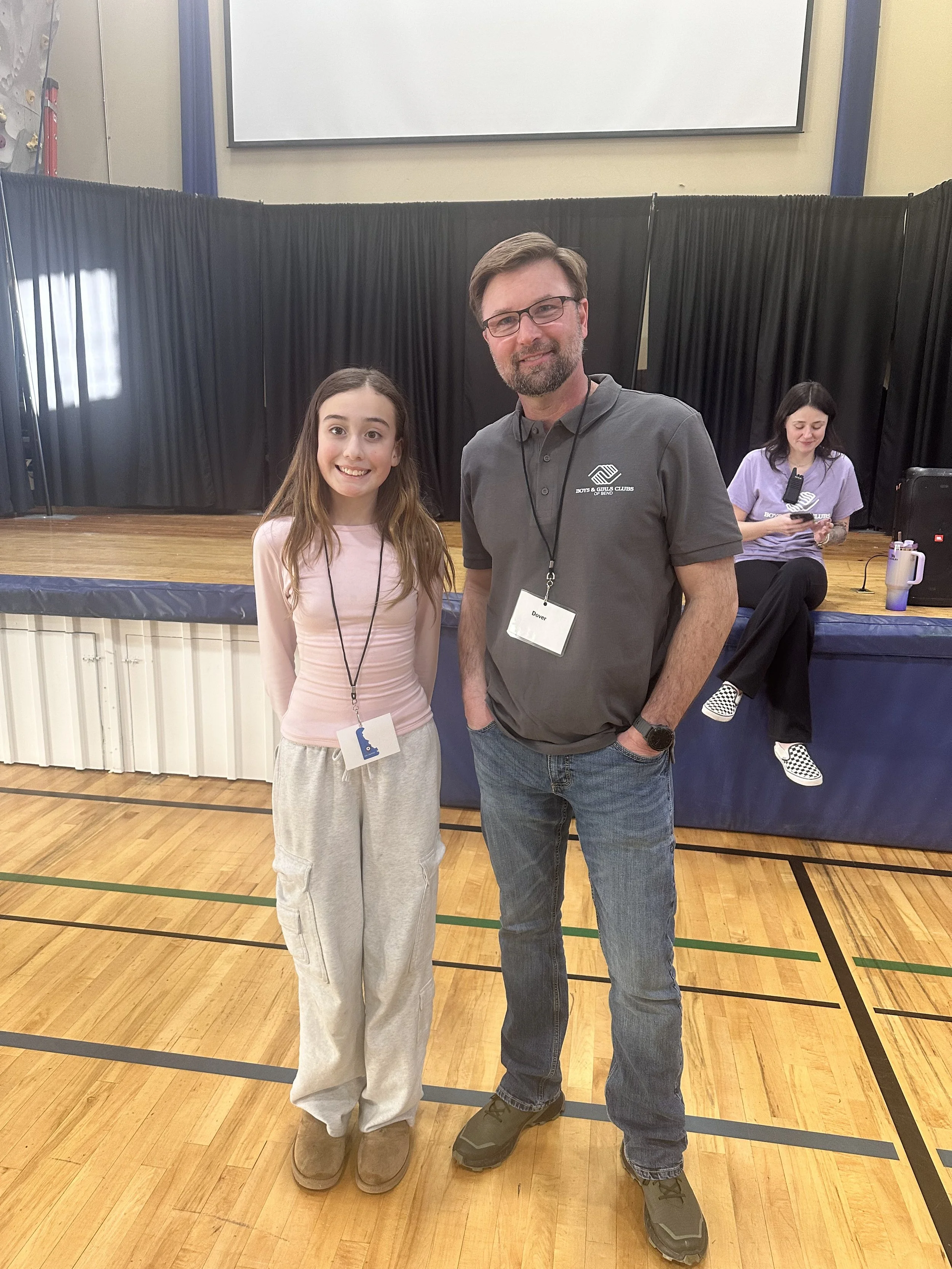 A young girl and adult man standing together inside a gymnasium, smiling at the camera. The girl is wearing light pink long-sleeve shirt and baggy gray sweatpants. The man is wearing glasses, a gray polo shirt with a logo, and jeans. In the backgroun