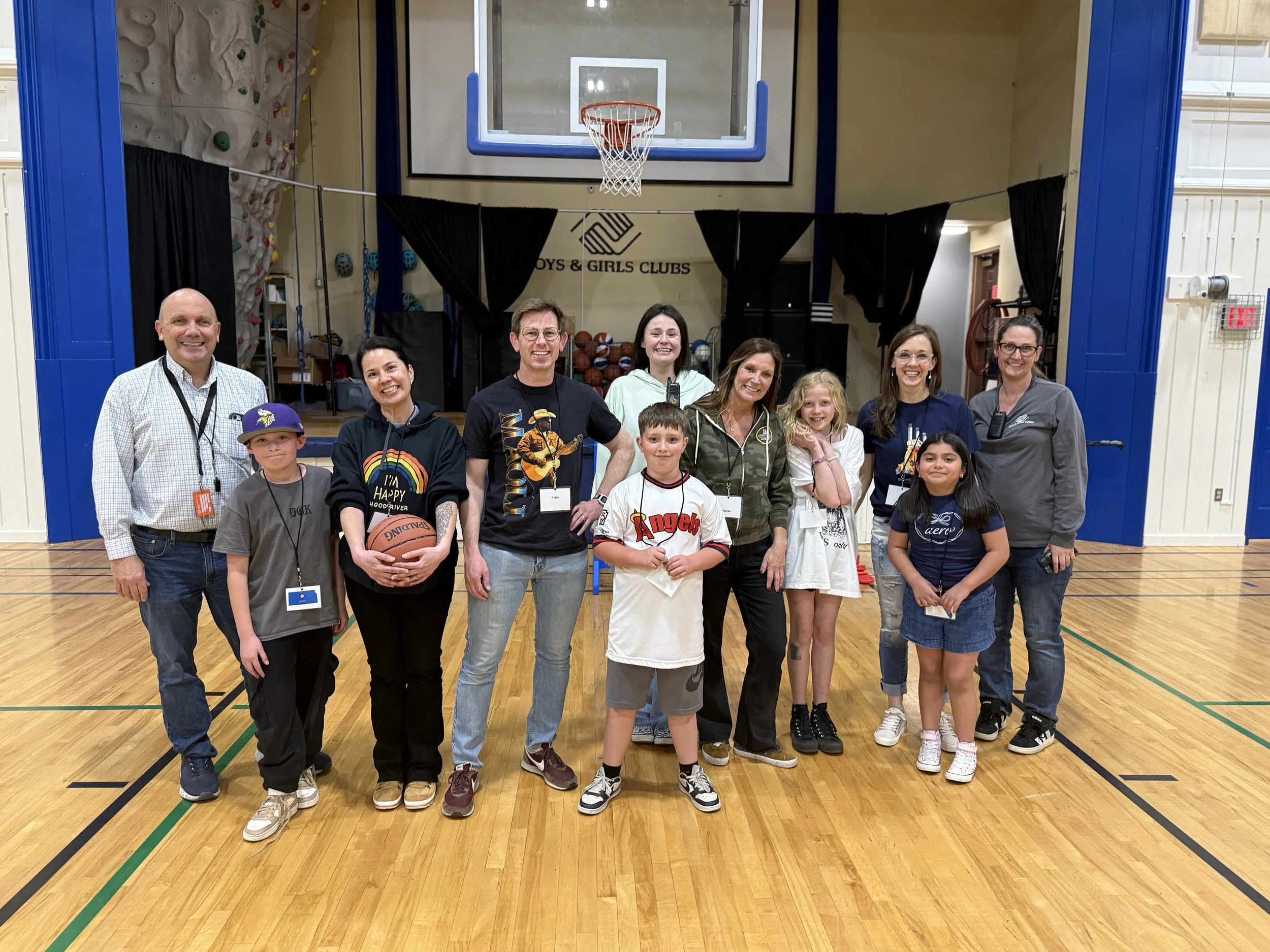 A group of adults and children smiling and posing together inside a gymnasium with a basketball hoop, a climbing wall, and black curtains in the background. The gymnasium has a wooden floor and the event appears to be related to Boys & Girls Clubs.