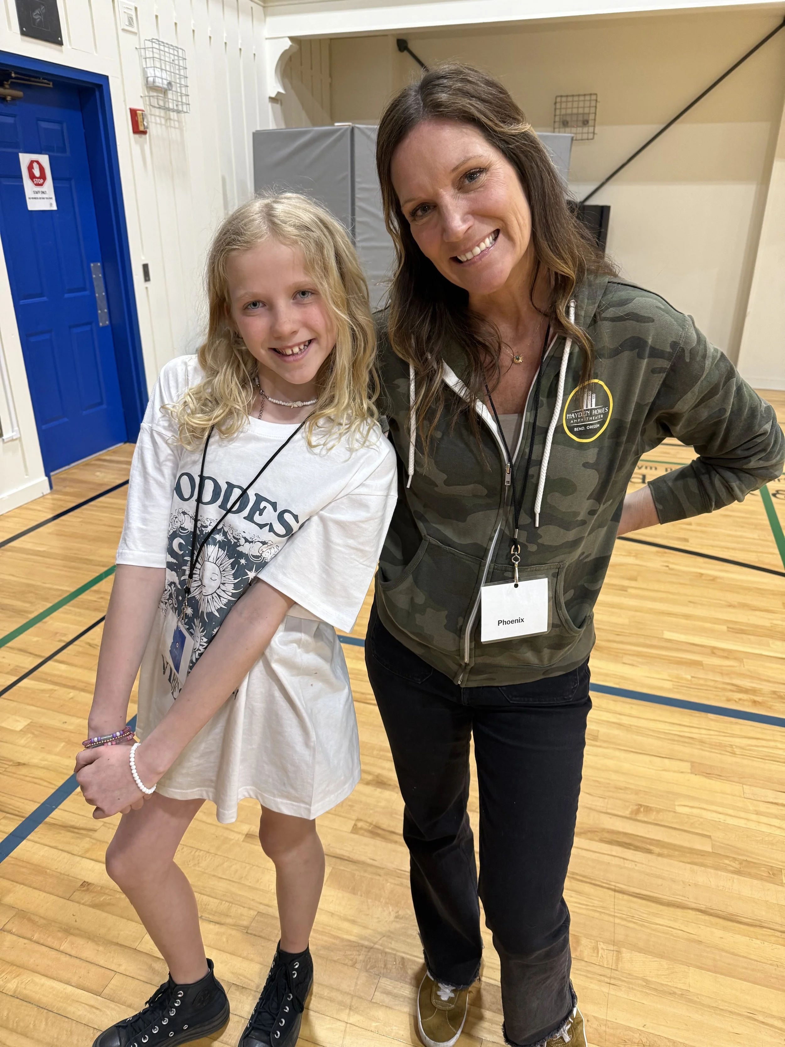 A woman and a young girl posing together indoors. The woman is wearing a camo jacket with a badge that says 'Hayden Homes' and a lanyard with a name tag that reads 'Phoenix'. The girl is wearing a white T-shirt with graphic prints, jewelry, and black