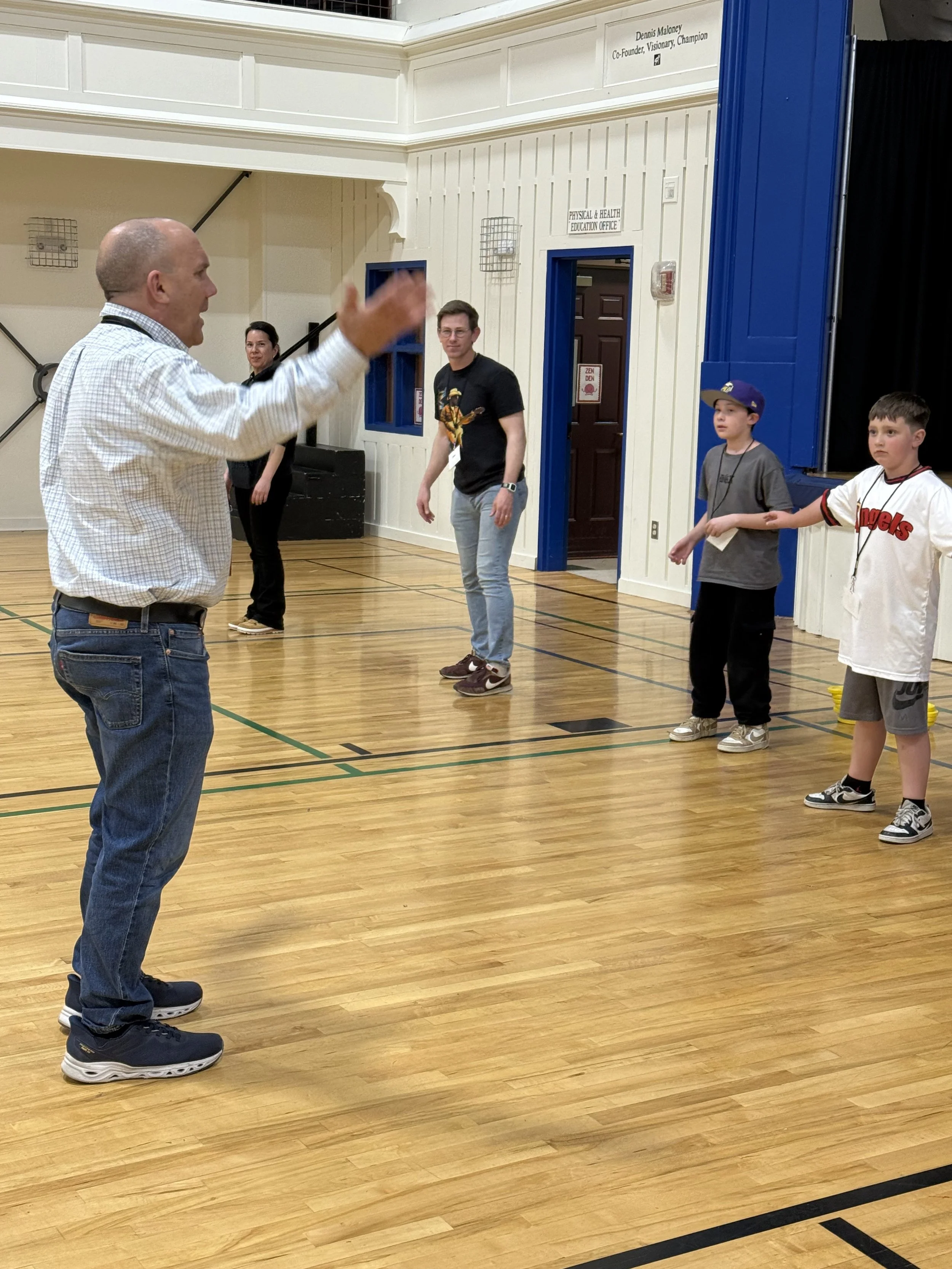 A man with a bald head and glasses speaking to children inside a gymnasium.