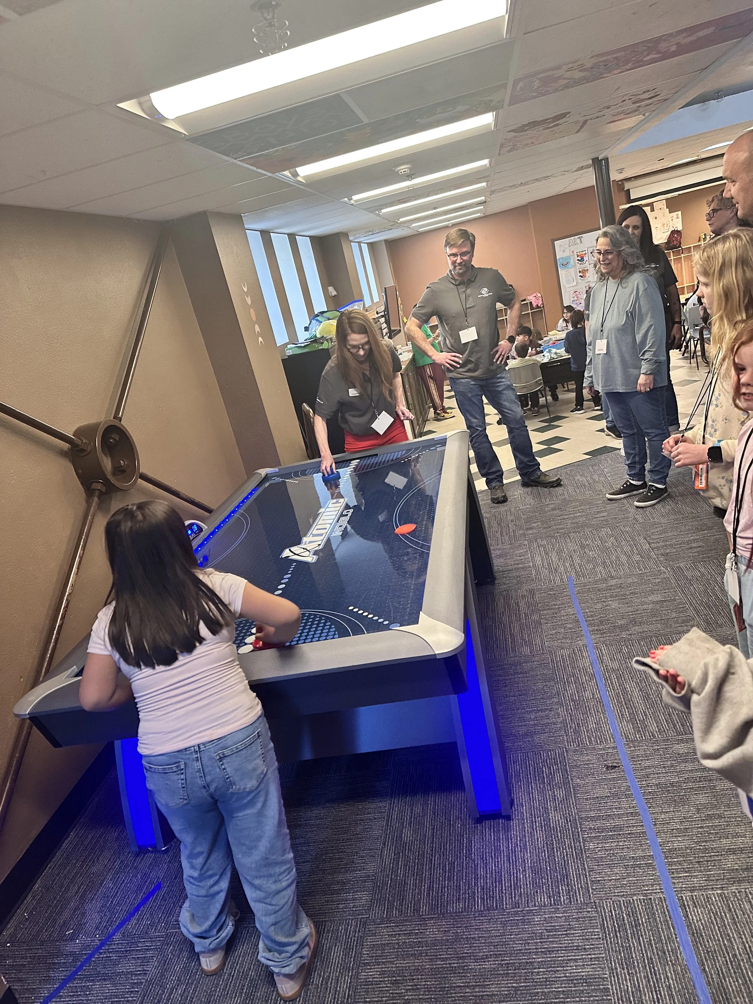 Children and adults gathered around an air hockey table, with children playing and adults observing in a room decorated with artwork on the walls.
