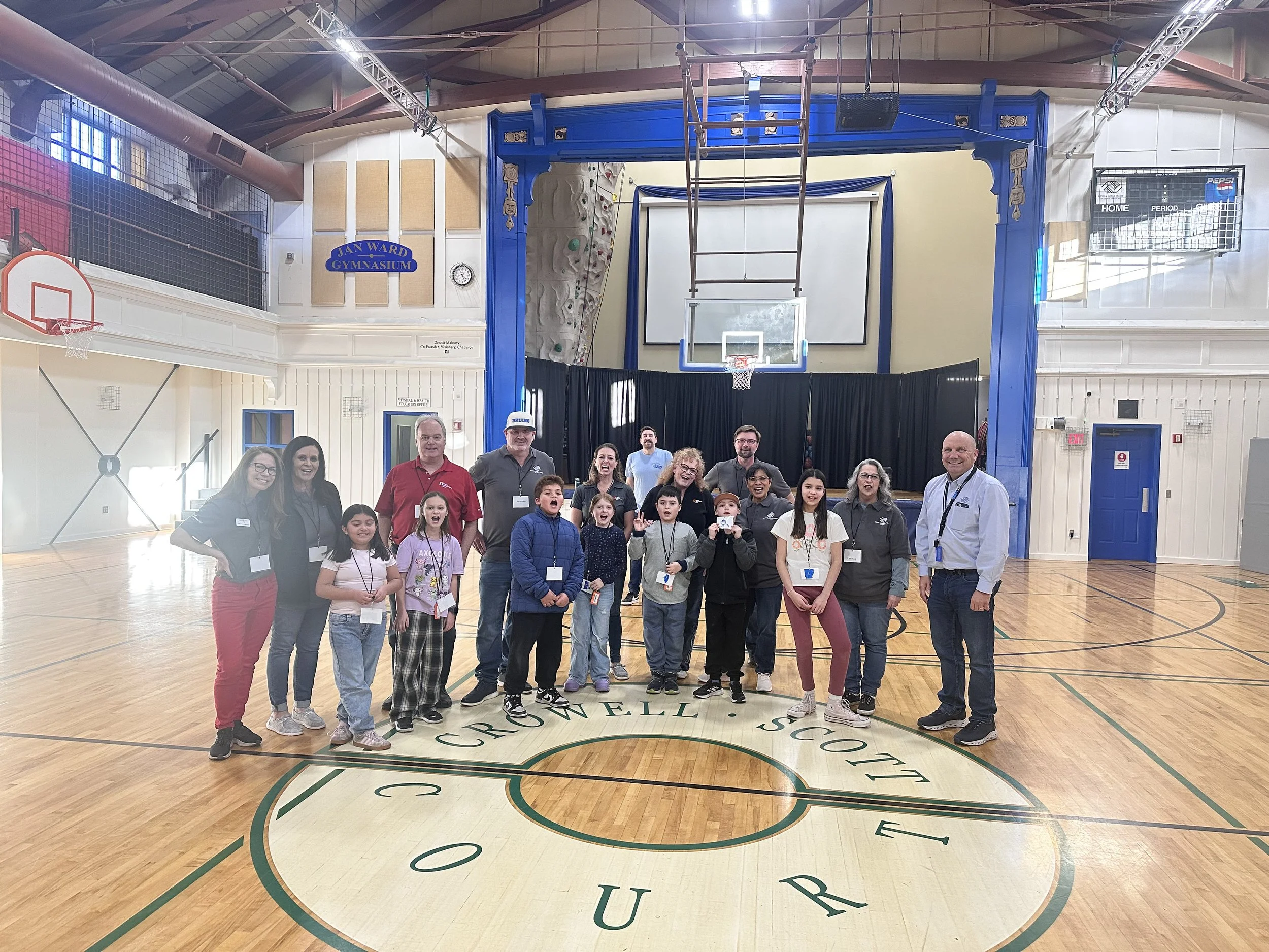 A group of children and adults posing for a photo in a gymnasium with a basketball hoop, a climbing wall, and a scoreboard. The gymnasium has a polished wooden floor with a large logo at the center.