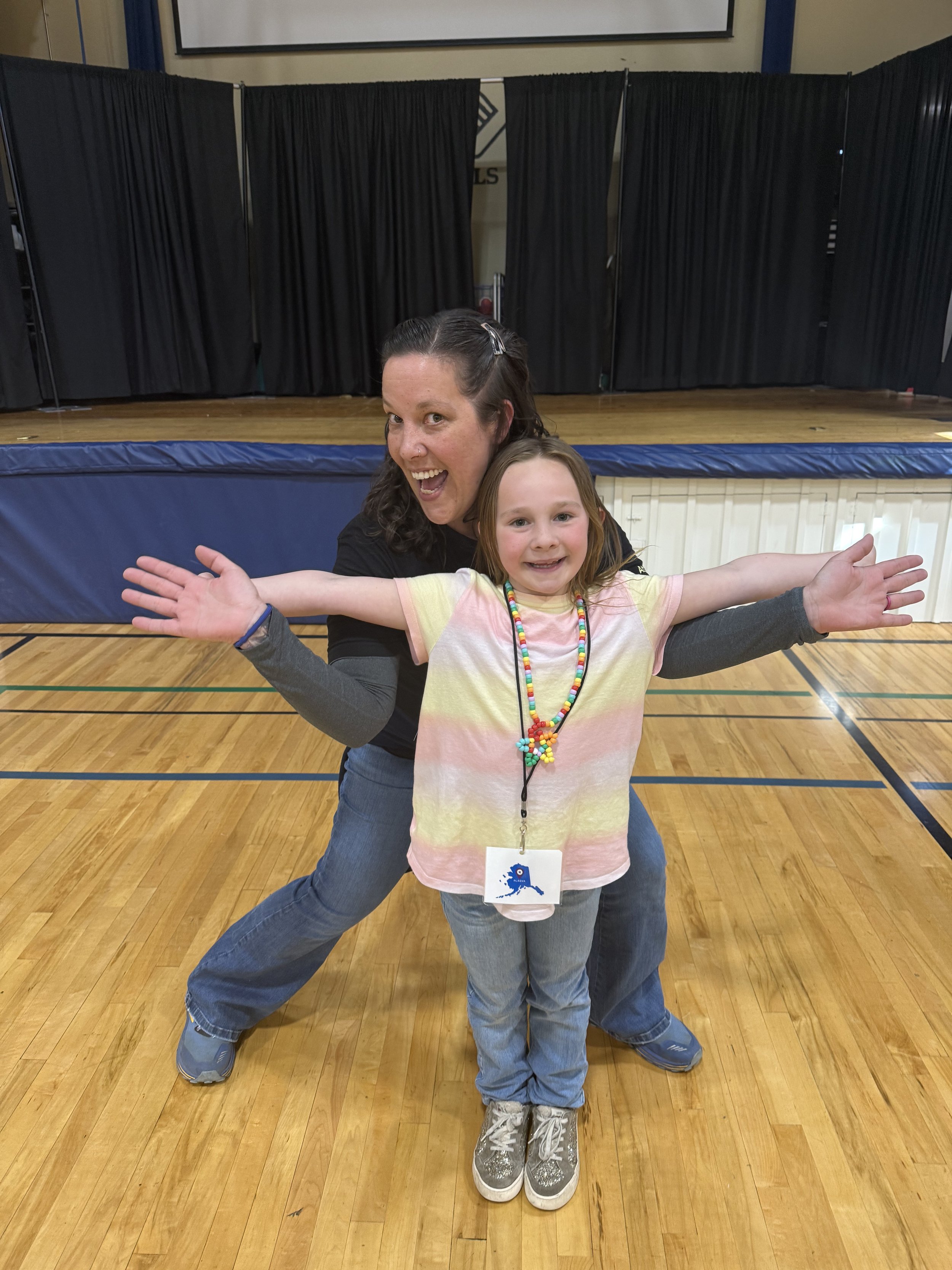 A woman and a young girl posing on a wooden gymnasium floor with black curtains and a stage in the background. The woman is wearing a black shirt and jeans, smiling with her arm around the girl. The girl is wearing a tie-dye shirt, colorful beaded ne