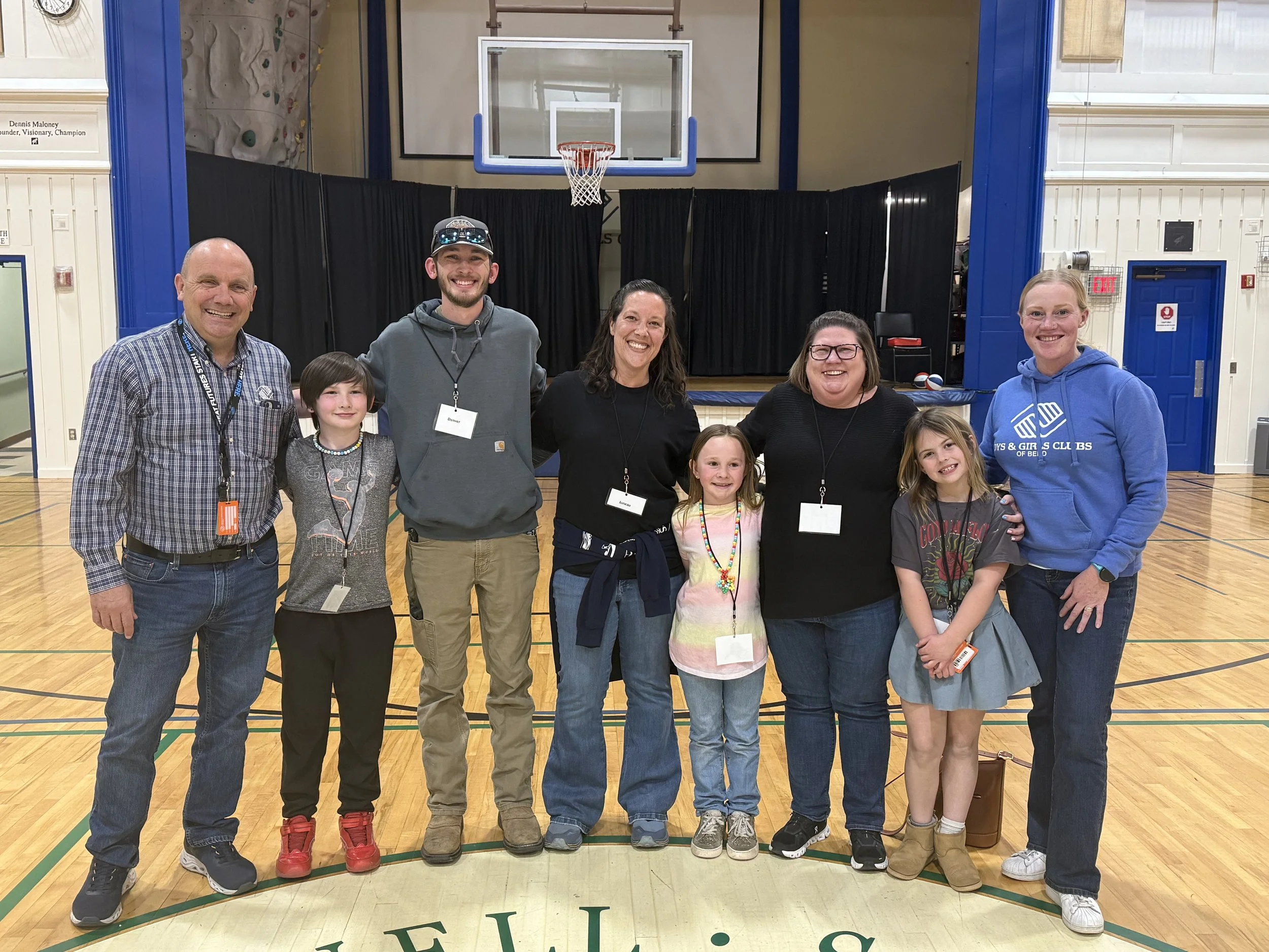 A diverse group of ten people, including children and adults, standing together in a gymnasium with a basketball hoop in the background. They are smiling and appear to be at a community or school event.