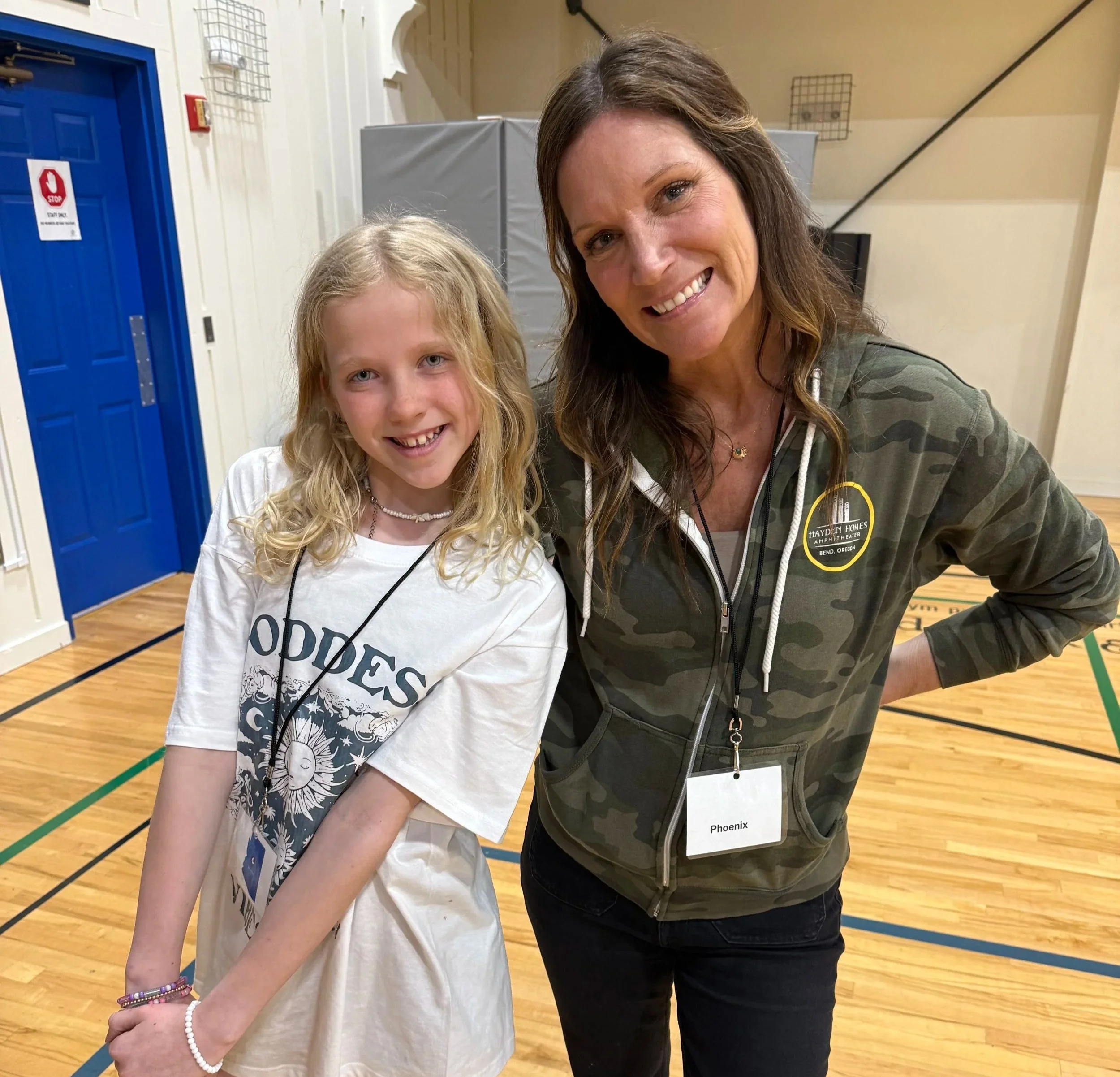 A young girl and a woman are taking a selfie in a gymnasium. The girl has blonde curly hair and is wearing a white t-shirt with a graphic design and beaded jewelry, smiling at the camera. The woman has long brown hair and is wearing a camouflage hoodie with a badge that says 'Phoenix' and a lanyard. They are both smiling and standing close together.