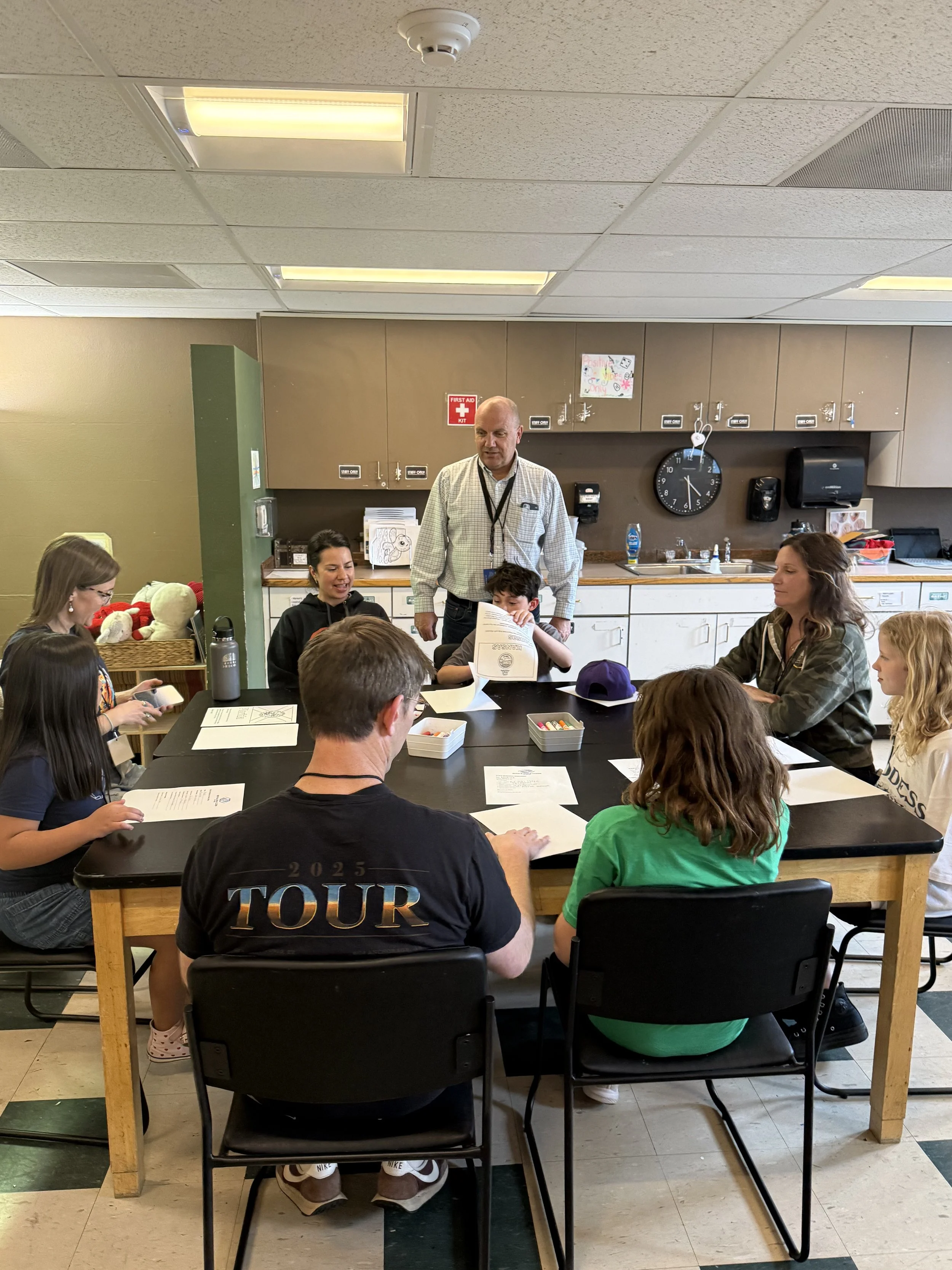 A group of children and adults sitting around a large table in a classroom, with some reading papers and a man standing in the center. The classroom has cabinets, a clock, a first aid kit, and children's artwork on the wall.