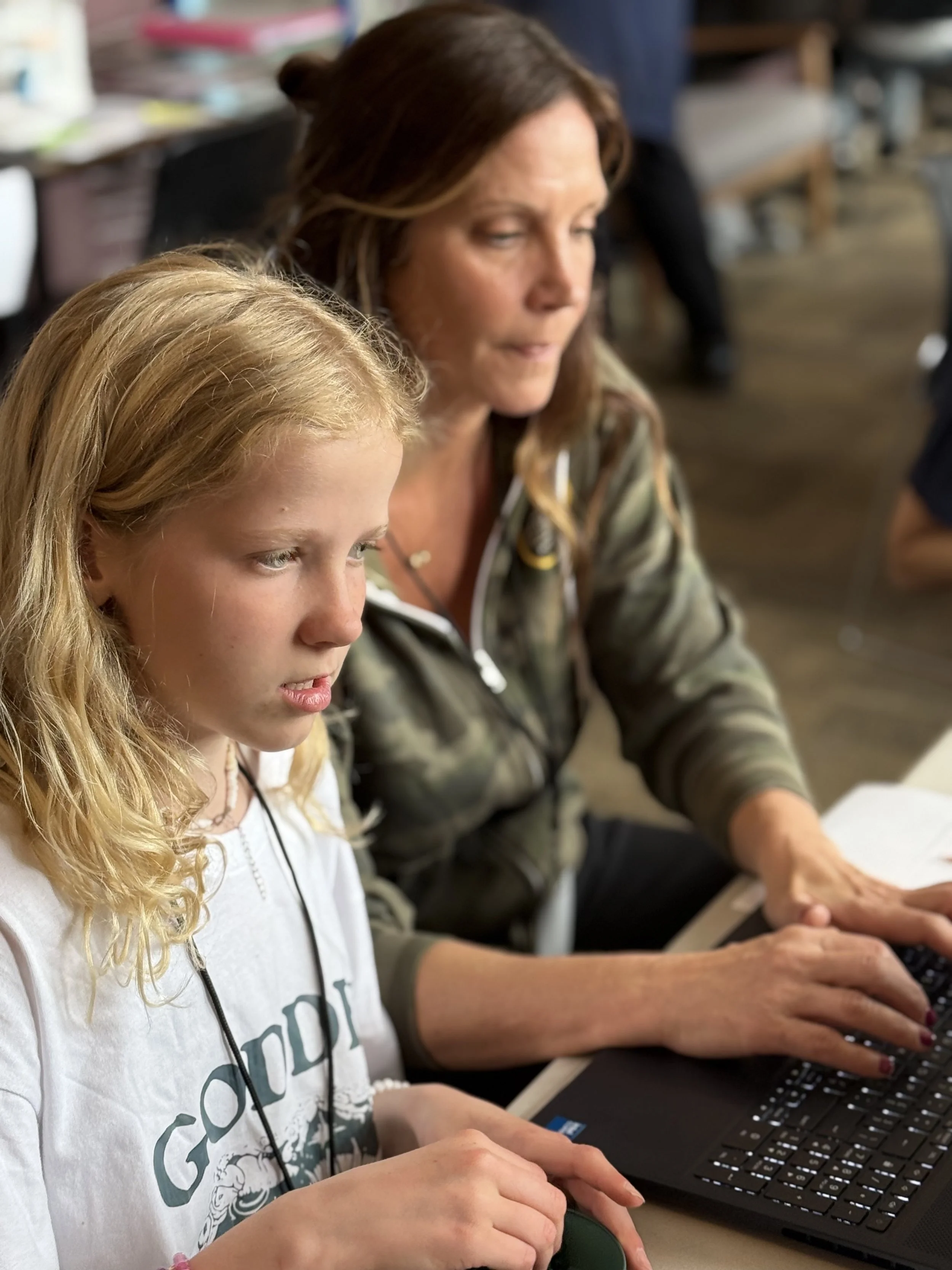 A woman and a girl are sitting at a table, working on a laptop. The woman has shoulder-length brown hair and the girl has long blonde hair. They are focused on the computer.