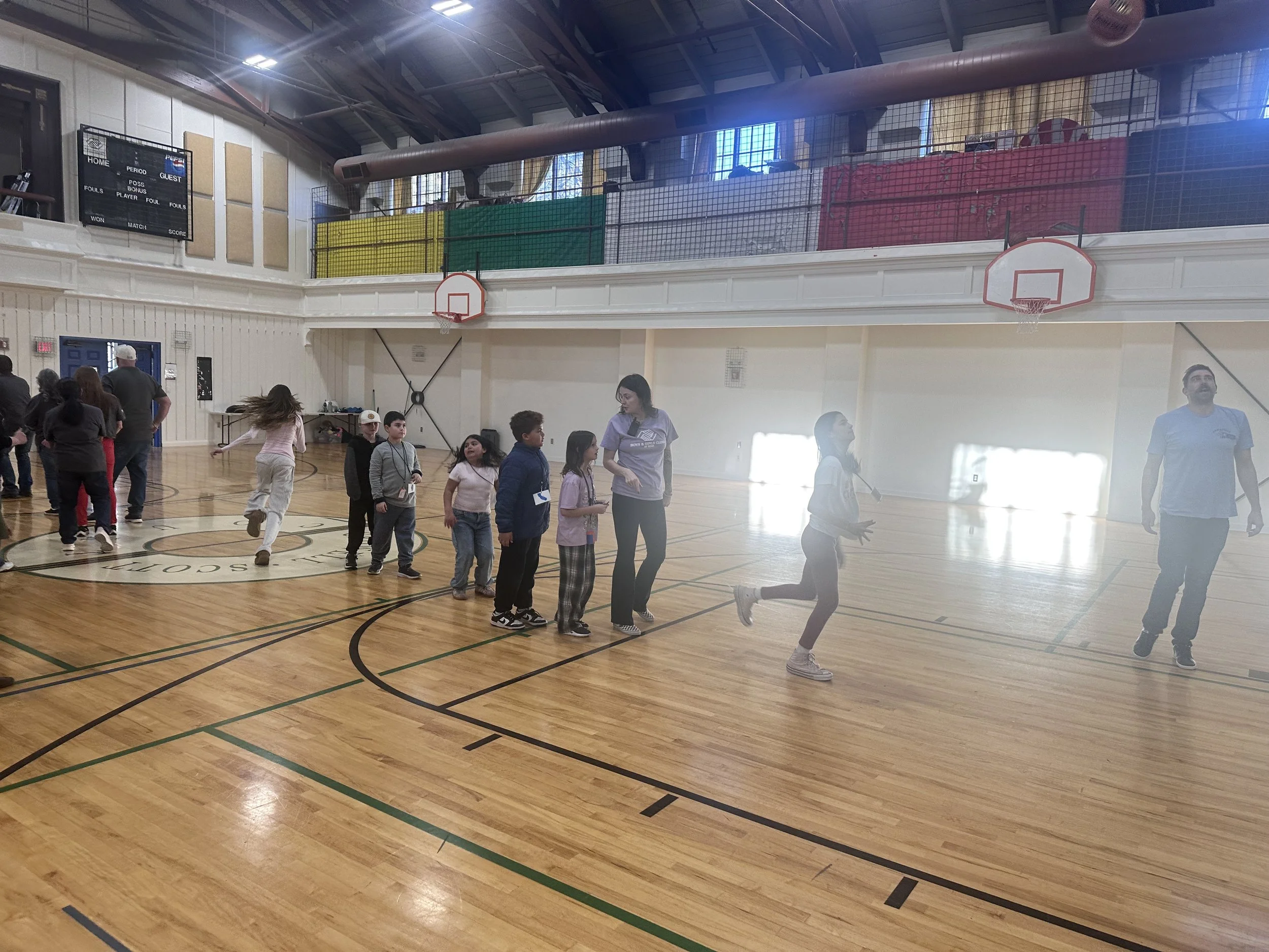 Children and adults inside a gymnasium with basketball hoops and a large scoreboard on the wall, engaging in activities and conversations.