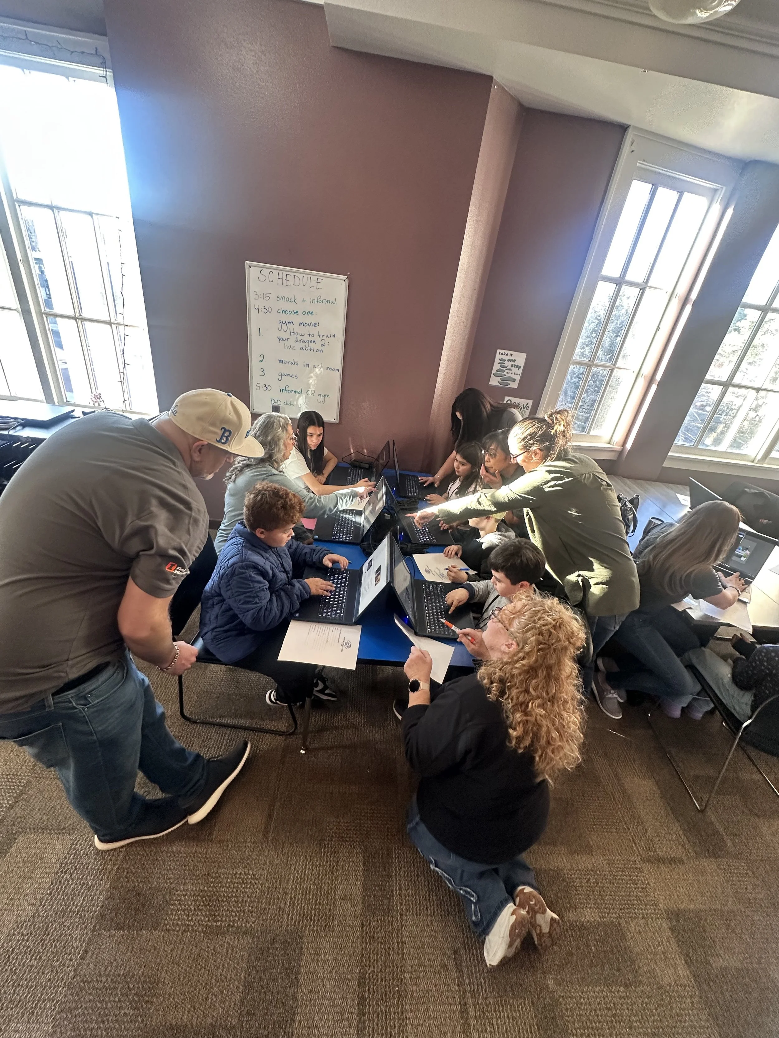 A group of children and adults gathered around laptops on a table, with some people sitting and others standing. Natural light from large windows illuminates the room. A whiteboard with a schedule and posters are on the wall.